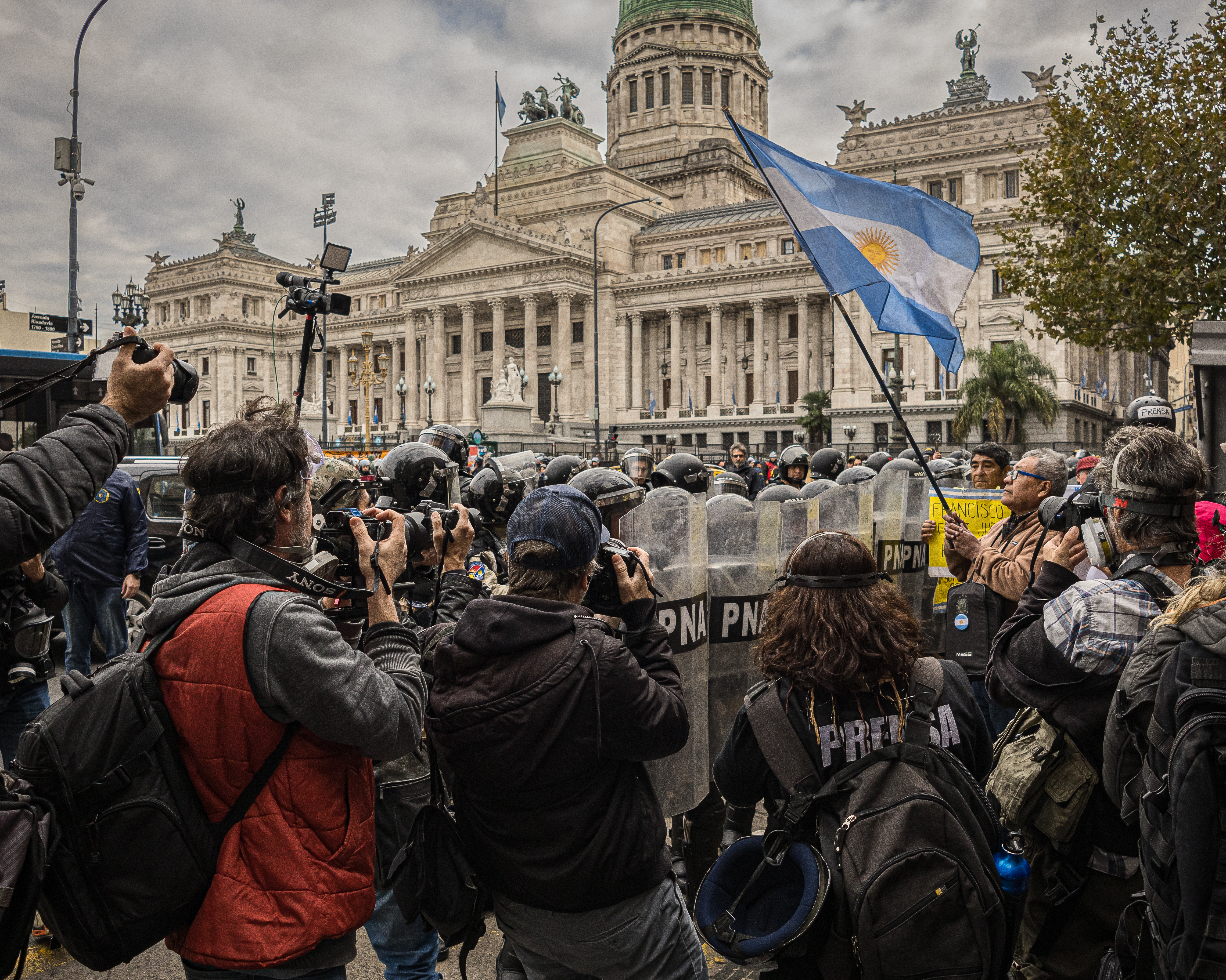 A wall of police officers in riot gear, with large plastic shields, stands near the Congress building while journalists take photographs and protesters demonstrate.