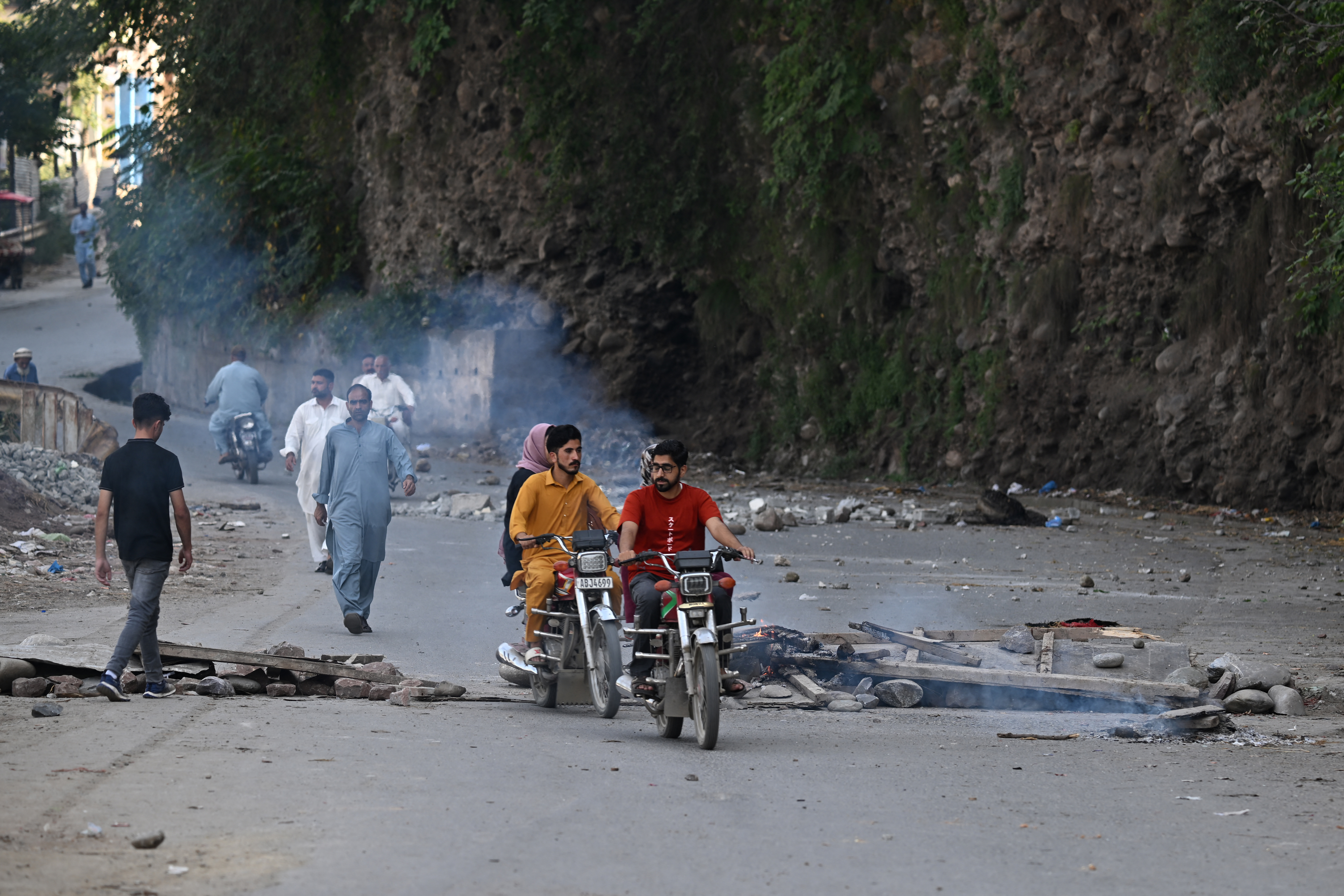 Commuters ride past burning logs of wood along a street blocked by Awami Action Committee (AAC) members during a demonstration in Muzaffarabad, capital of Pakistan-administered Kashmir on October 1, 2025, demanding structural reforms and political and economic rights.
