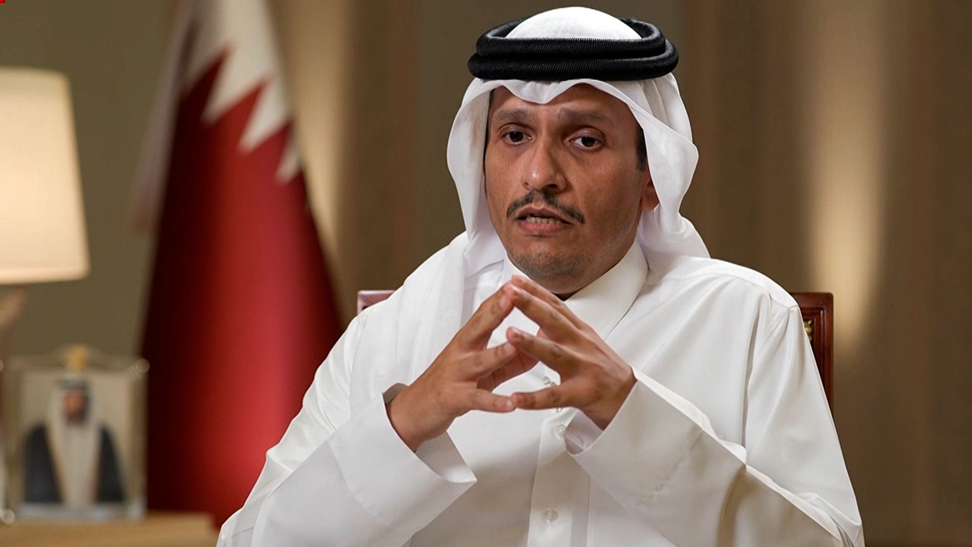 Mohammed bin Abdulrahman Al Thani, Prime Minister of Qatar, wearing a white thobe and ghutra, with hands clasped, in front of a Qatari flag.