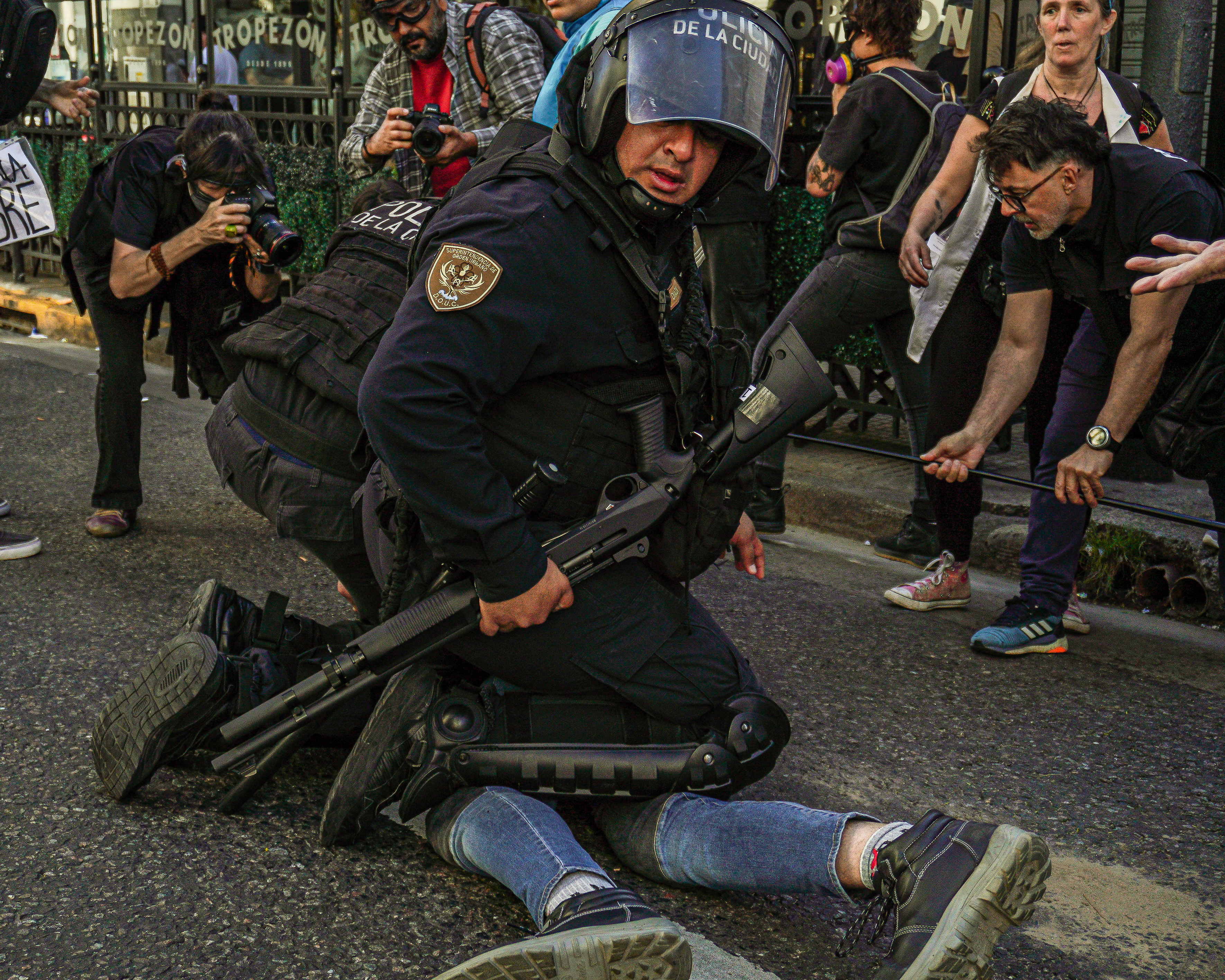 A police officer in riot gear kneels on a protester, whose feet are the only part visible beneath the officer's body.