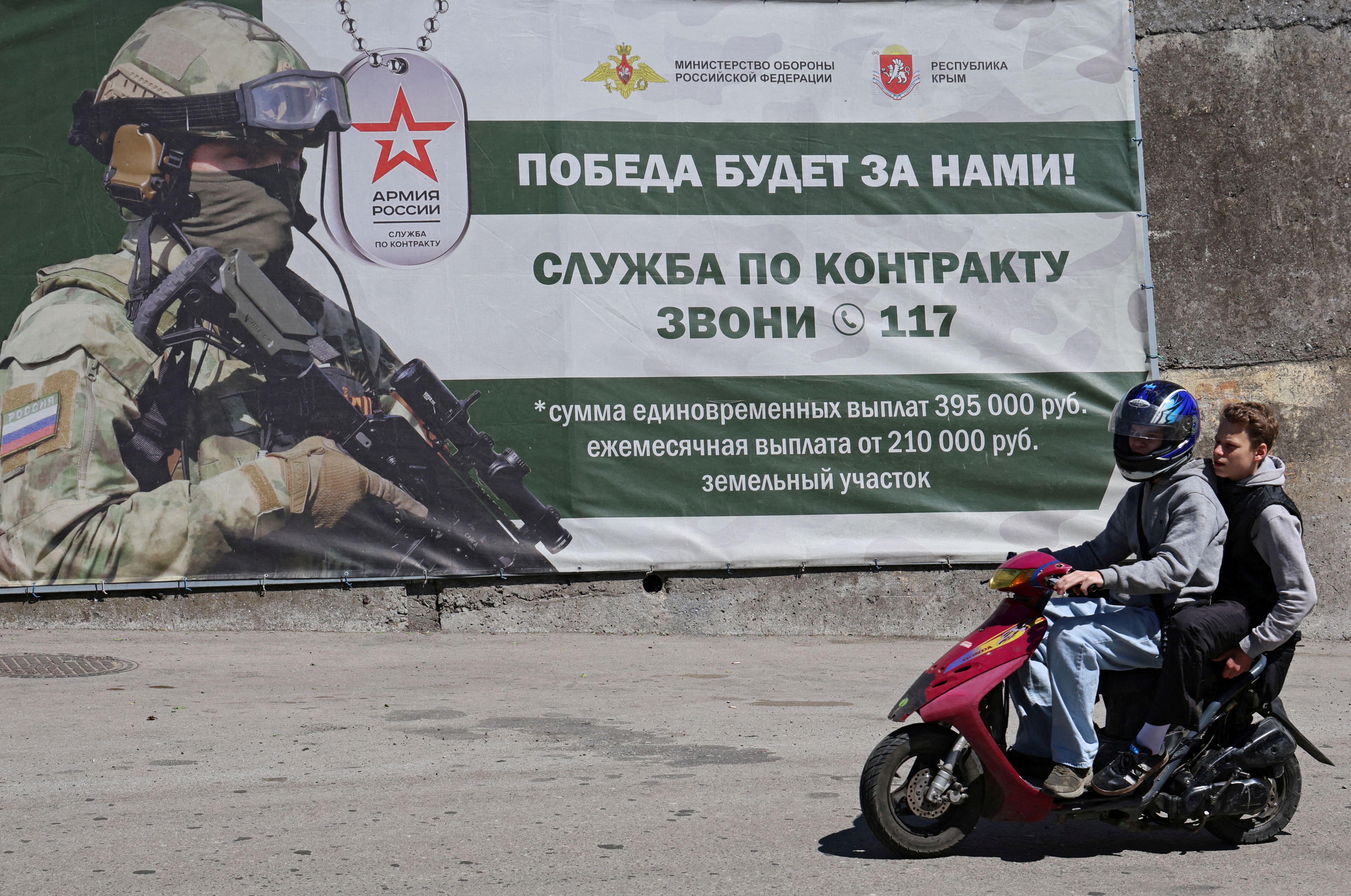 People ride a moped past a banner promoting military service in the Russian Armed Forces.