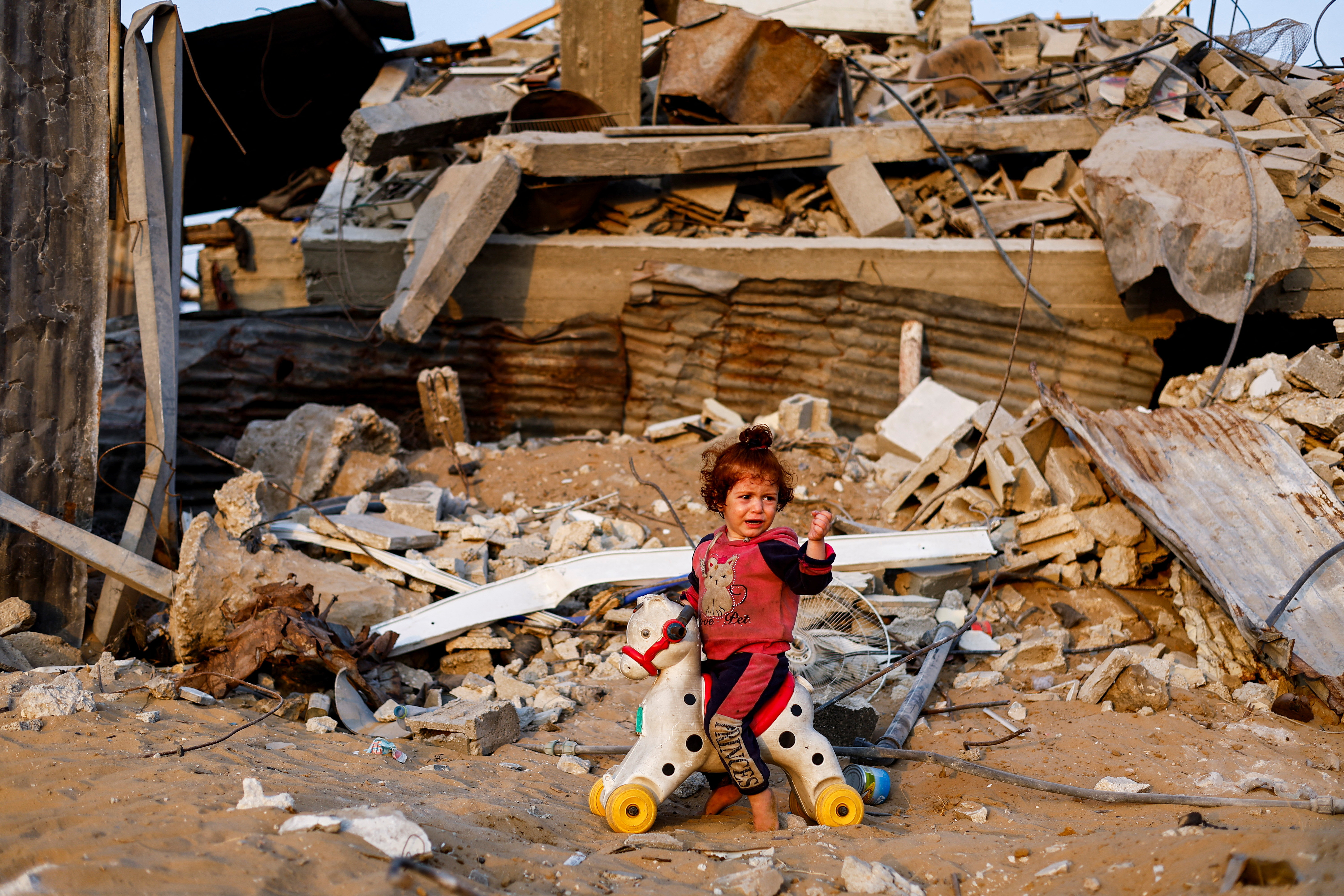 A Palestinian child sits on a toy horse amid the rubble of destroyed buildings.