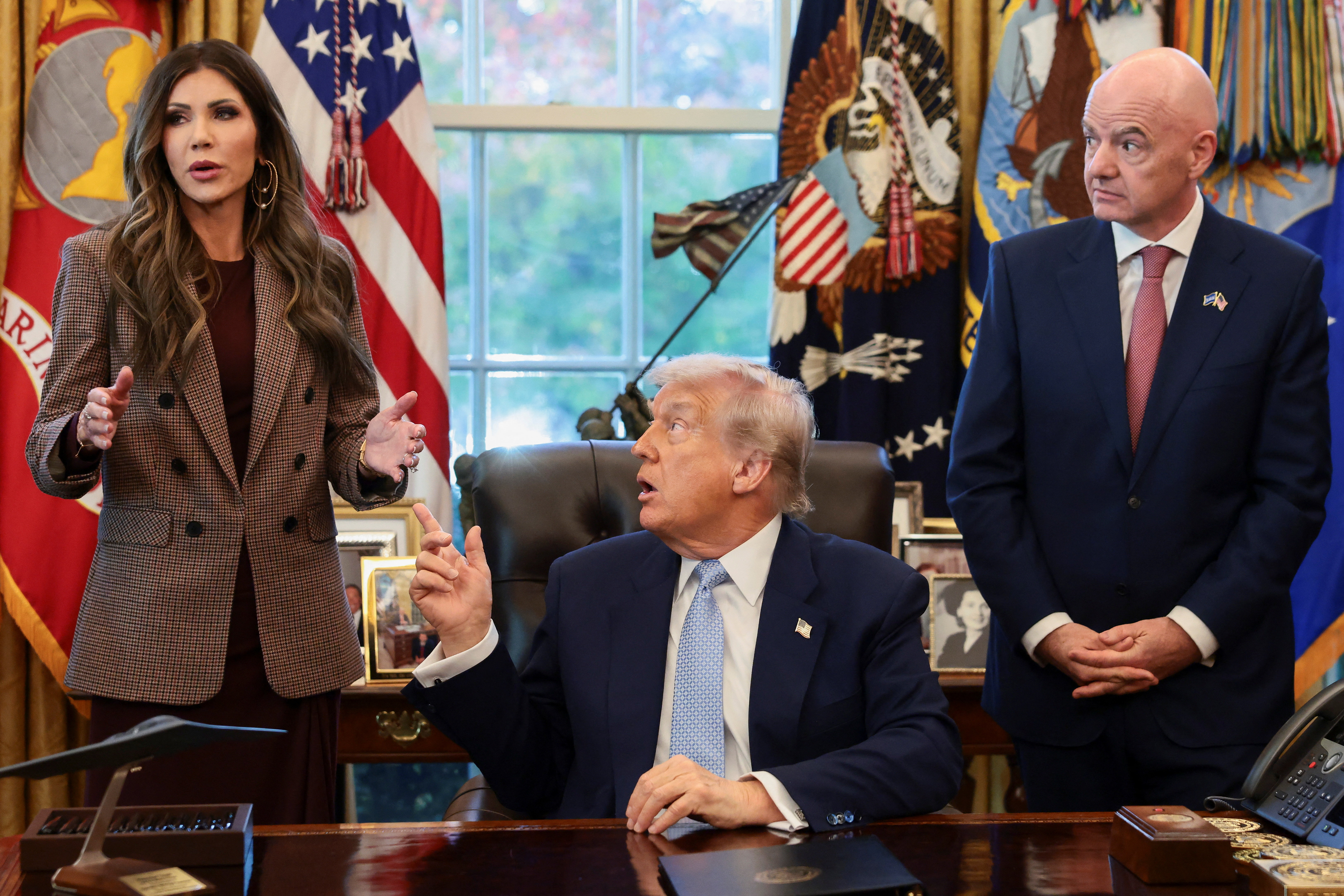 US President Donald Trump sits while FIFA President Gianni Infantino and US Homeland Security Secretary Kristi Noem stand by his side.