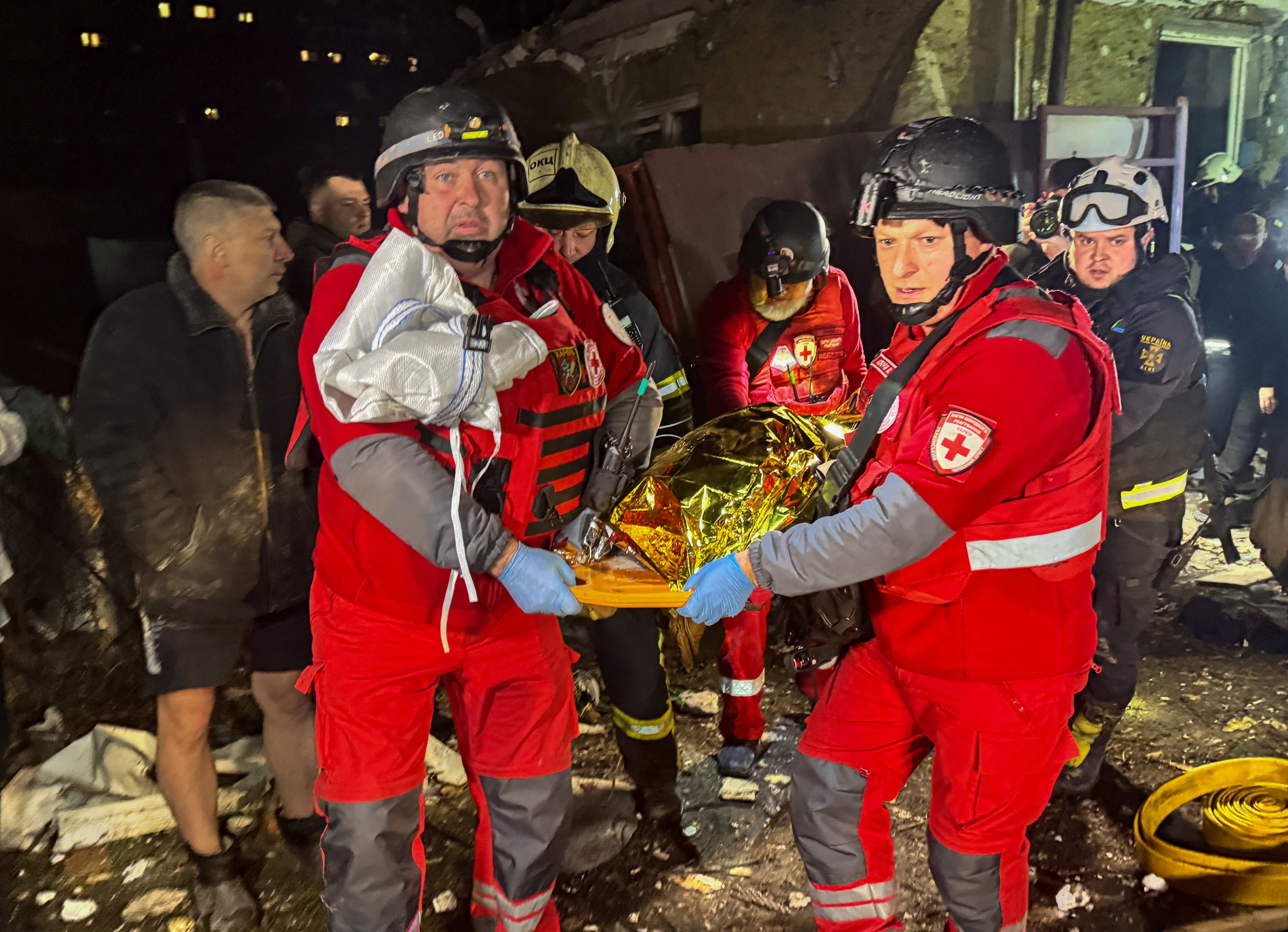 men in red emergency uniforms carry a body wrapped in a gold heat blanket on a stretcher at night