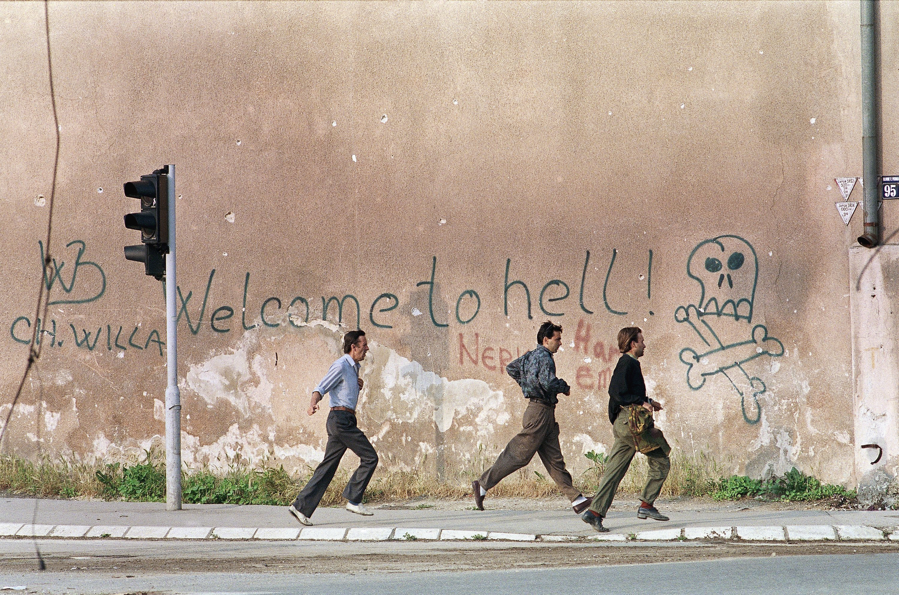 Three men run for cover past graffiti reading: "Welcome To Hell" as sniper fire rings out along Sarajevo's notorious "sniper alley"