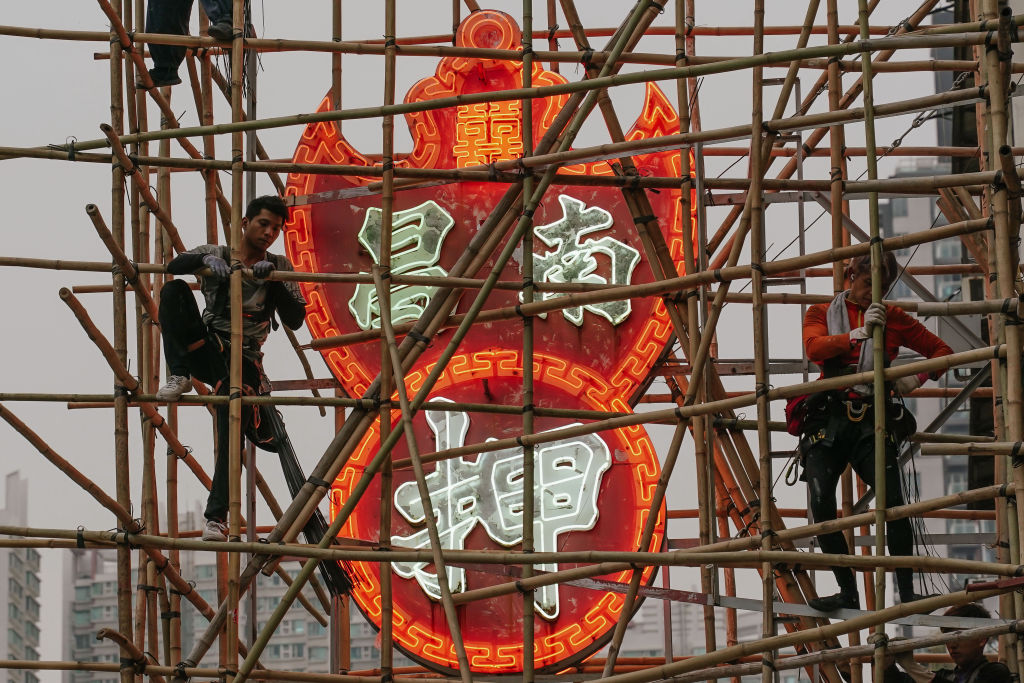workers stand on bamboo next to a red neon sign