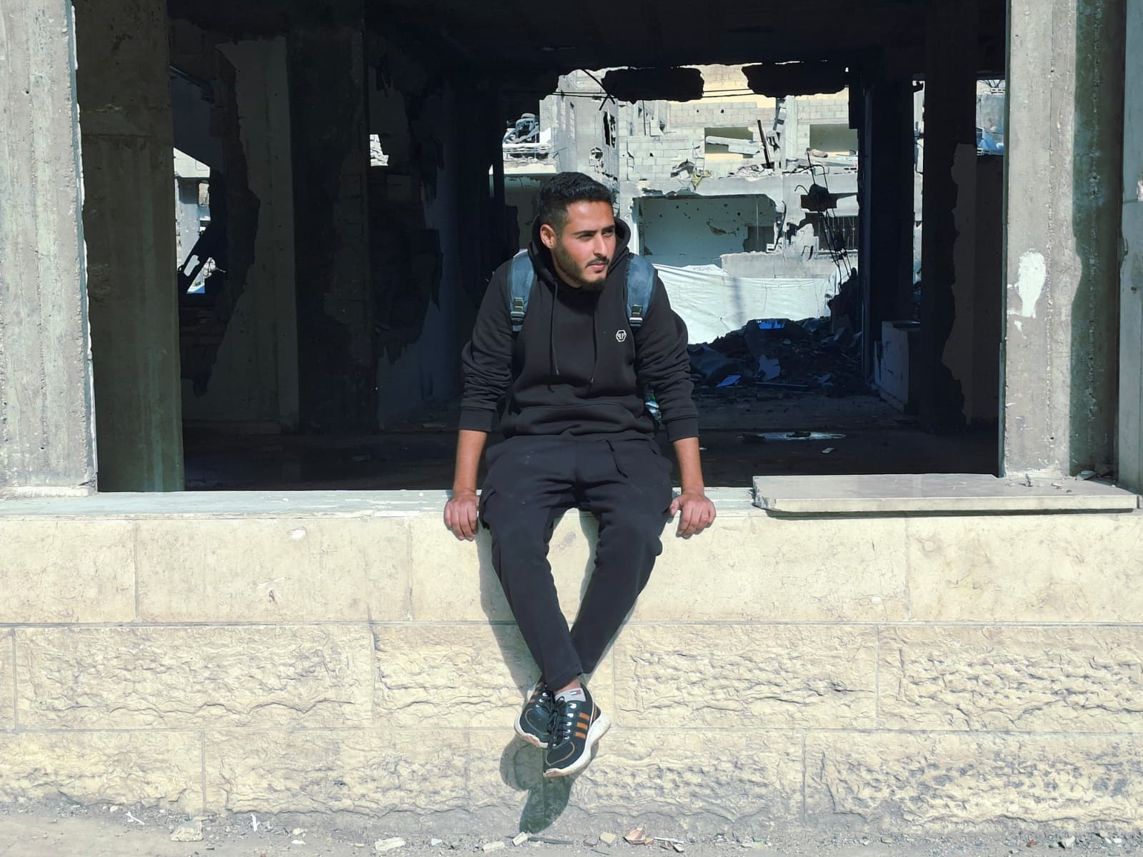 A young man sitting on a short wall and behind him is a scene of destruction