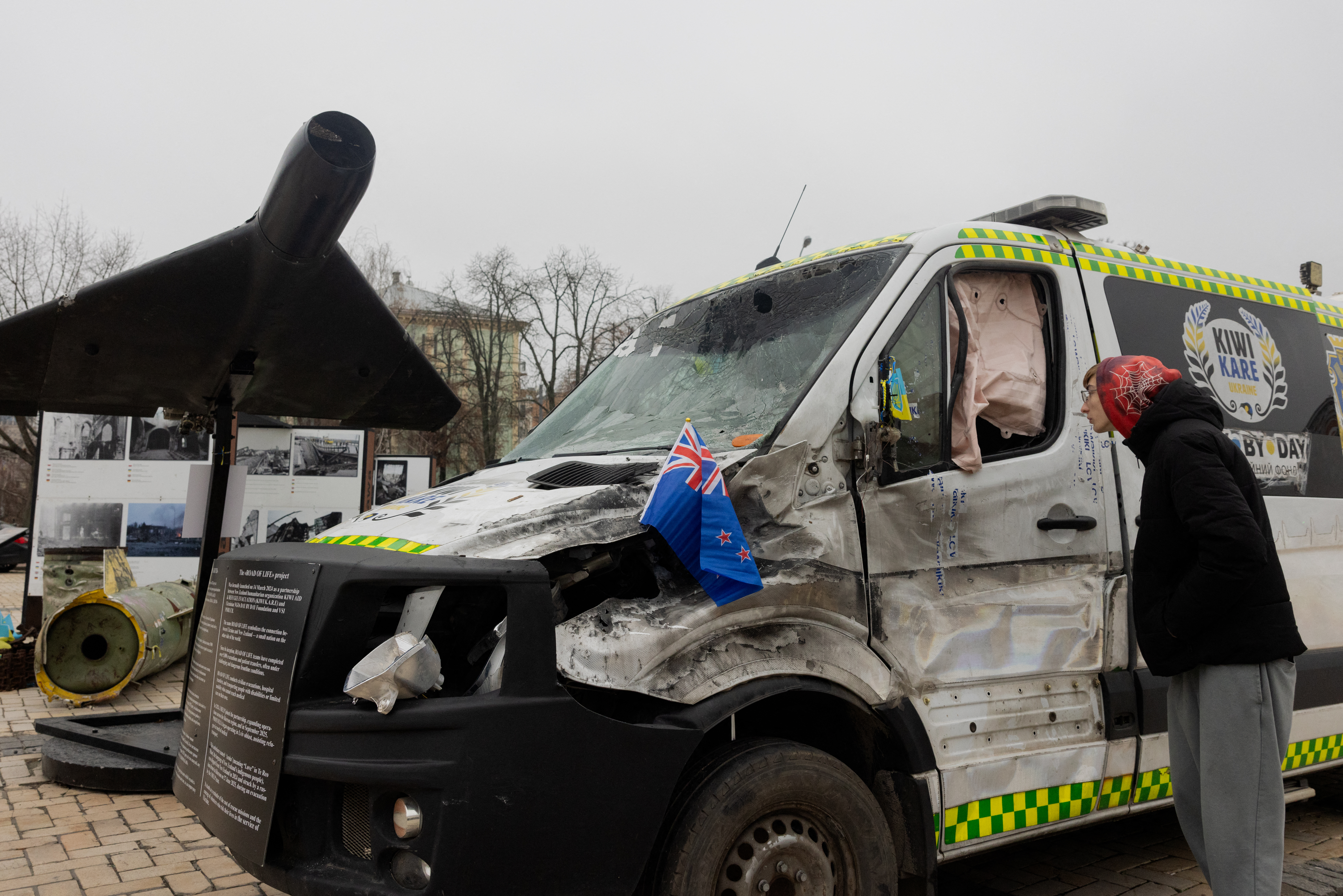 A man looks at a destroyed Kiwi K.A.R.E ambulance launched as part of a humanitarian project of "The Road of Life" between New Zealand and Ukraine, which was struck by a Russian drone during an evacuation mission in June 2025, next to an Iranian-designed Shahed 136 (Geranium-2) drone of Russian Army in an open-air exhibition of destroyed Russian military equipment on Mykhailivska Square (Saint Michael's Square) in Kyiv on December 9, 2025, amid the Russian invasion of Ukraine. (Photo by Tetiana DZHAFAROVA / AFP)