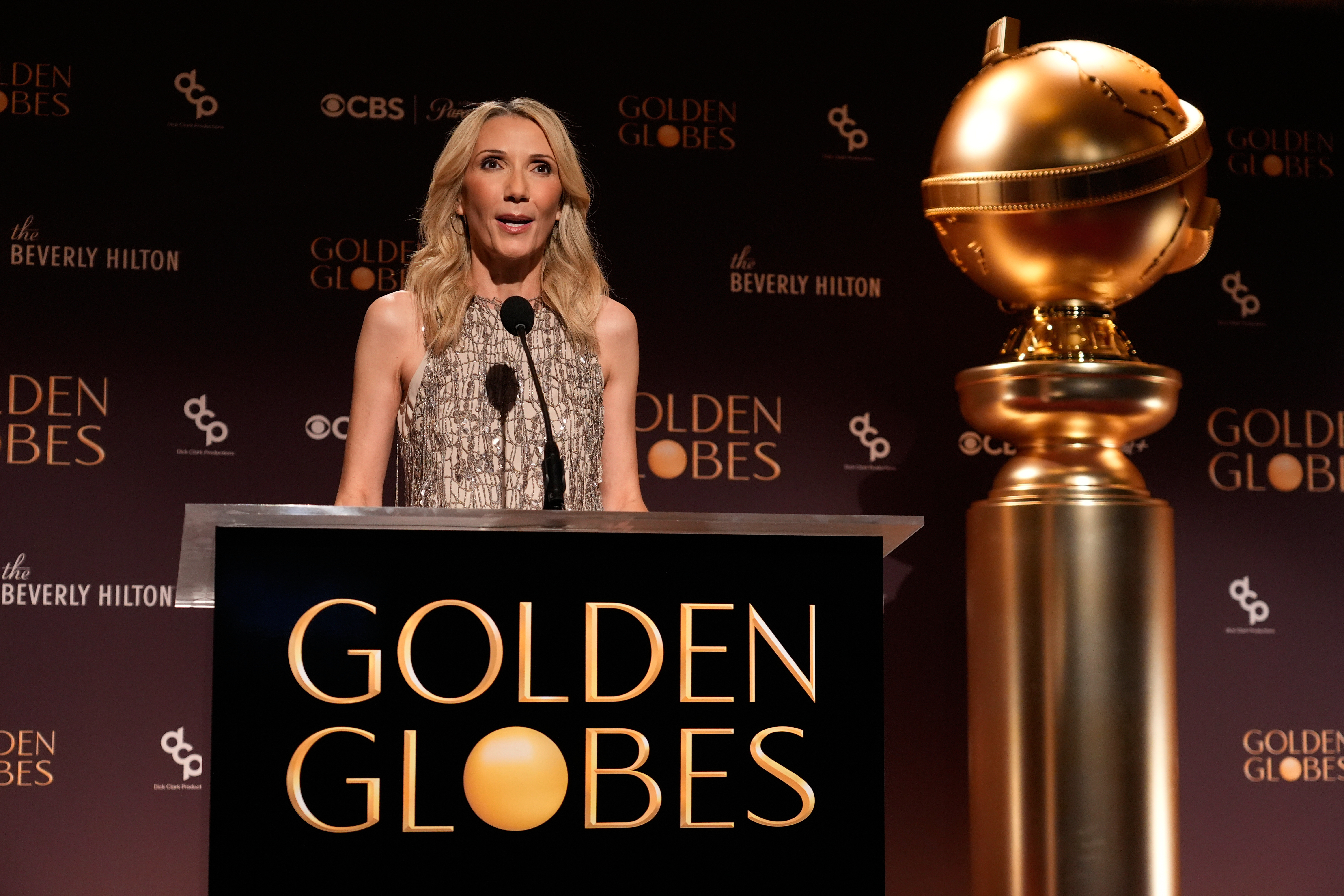 Golden Globes president Helen Hoenhe at a Golden Globes-branded podium, next to a large version of the award.
