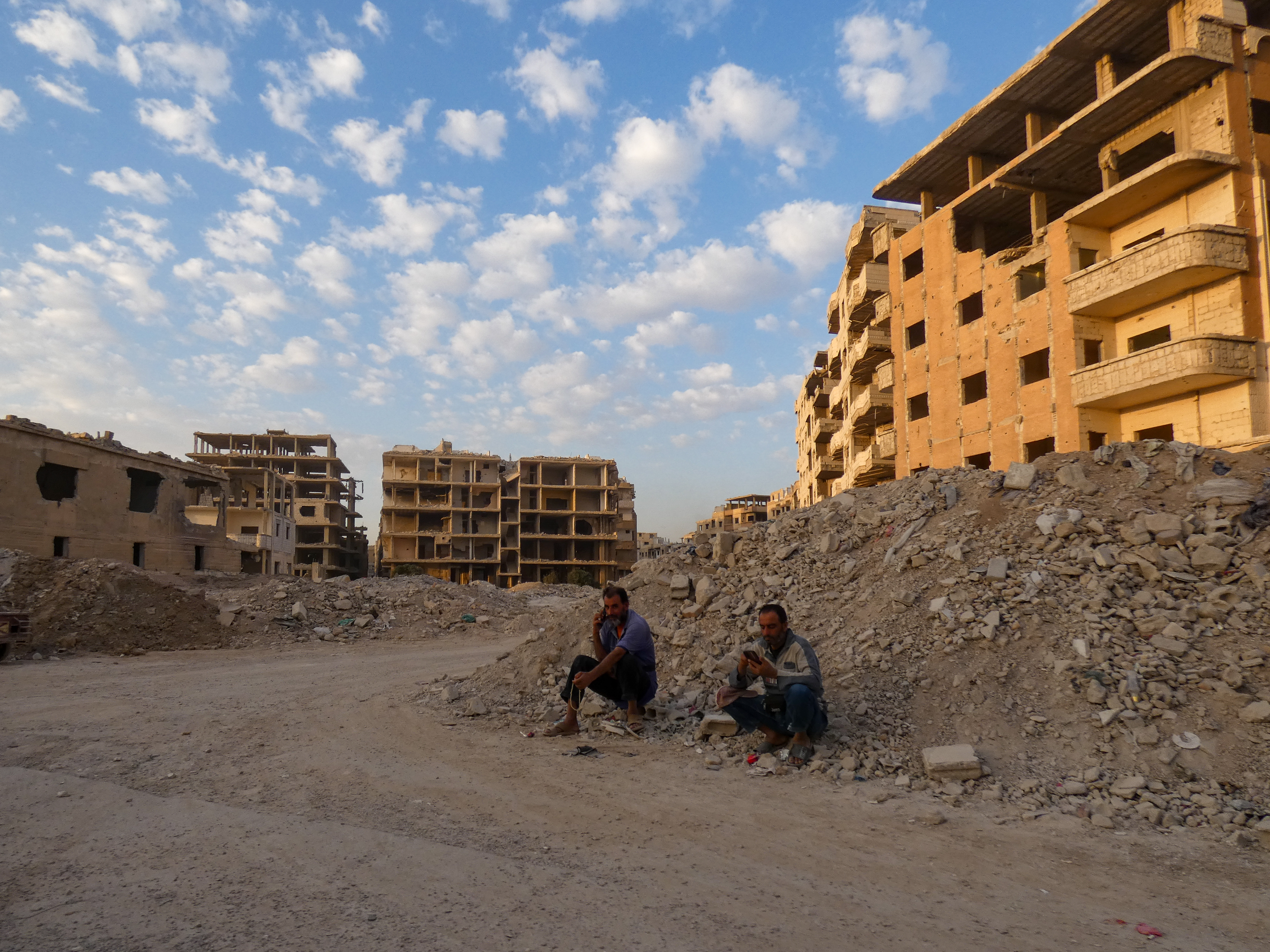 Two men sit among the rubble in Tadamon with buildings in the background.