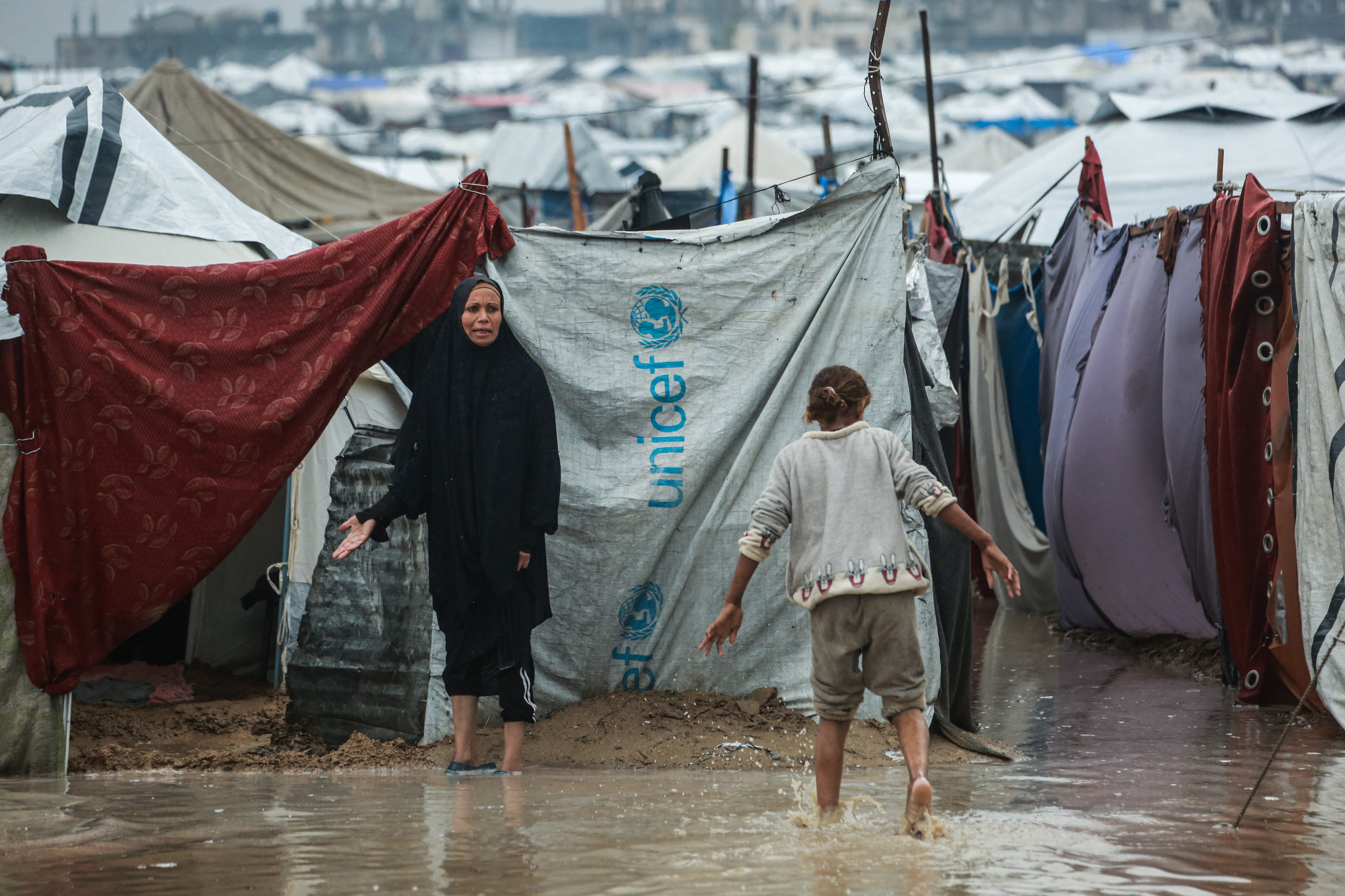 A woman gestures as a girl walks through a pool of water at a makeshift camp housing displaced Palestinians in Deir al-Balah, in the central Gaza Strip, on December 11, 2025.