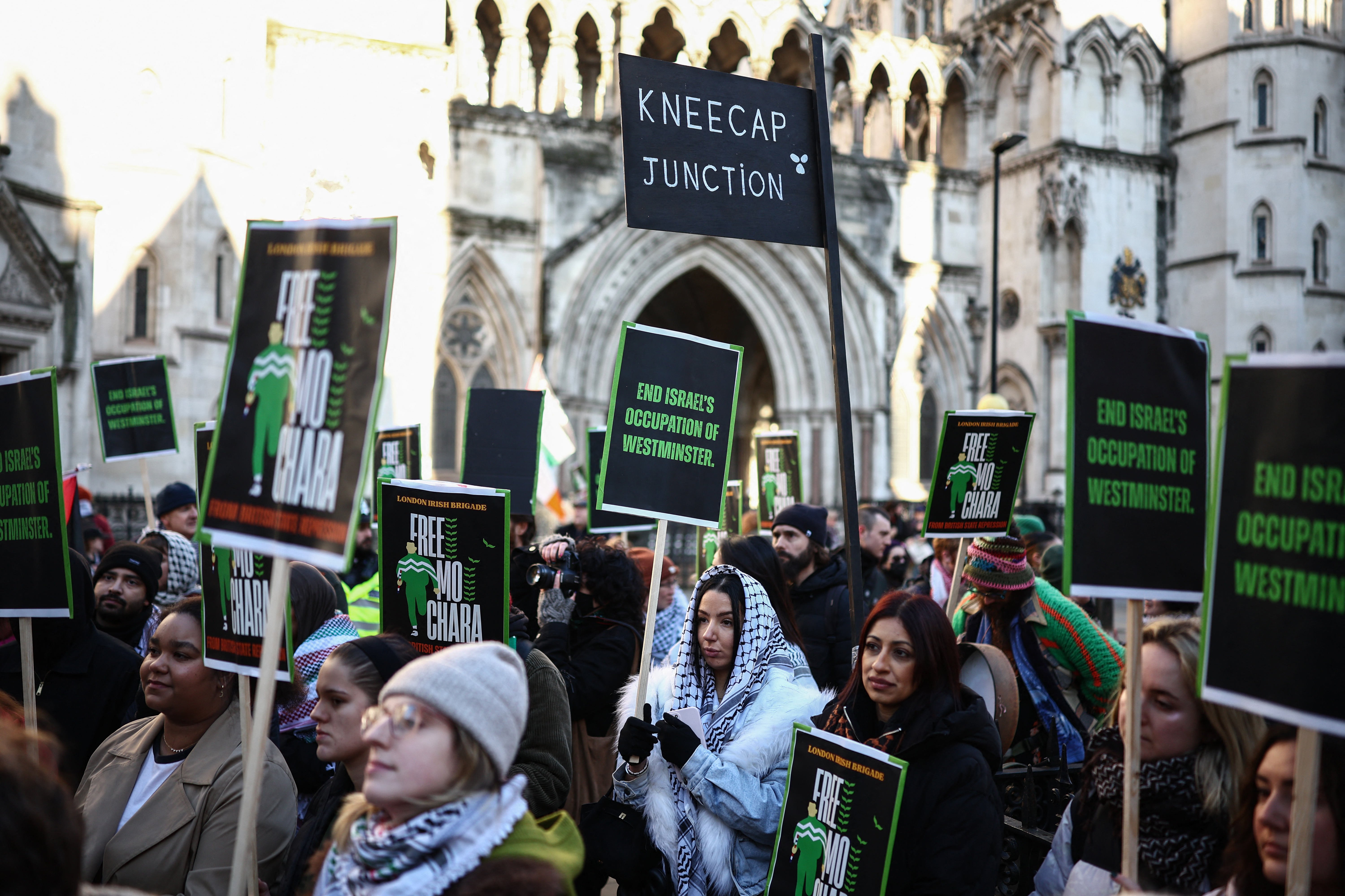Supporters of Irish rap group Kneecap band member, Liam O'Hanna, who performs under the stage name Mo Chara, hold placards as they gather outside the Royal Courts of Justice ahead of the singer's arrival in London on January 14, 2026.