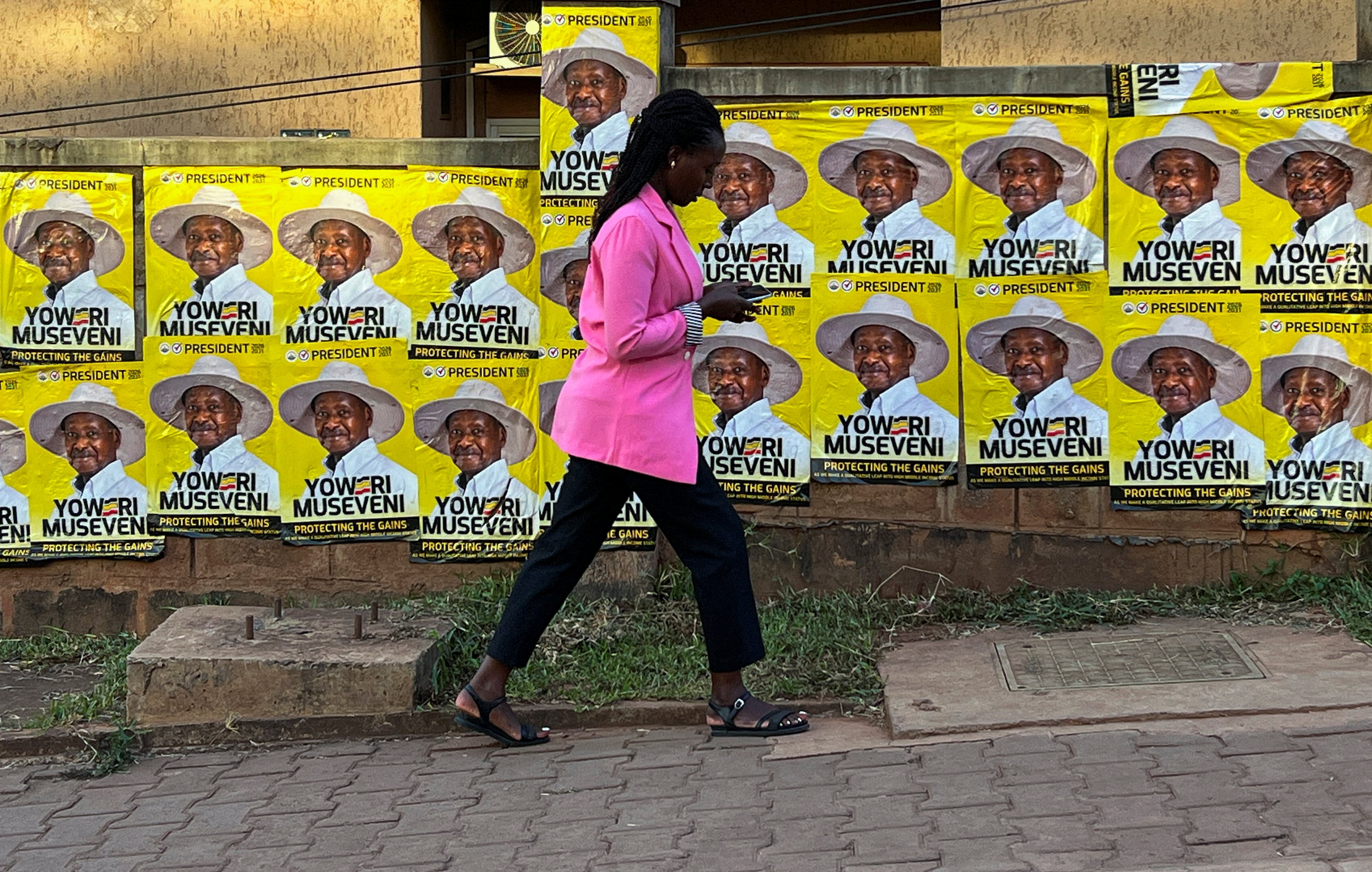 A woman uses a mobile phone as she walks past campaign posters of Yoweri Museveni, Uganda's President and presidential candidate of the ruling National Resistance Movement (NRM), ahead of the general election in Kampala, Uganda, January 14, 2026. REUTERS/Thomas Mukoya