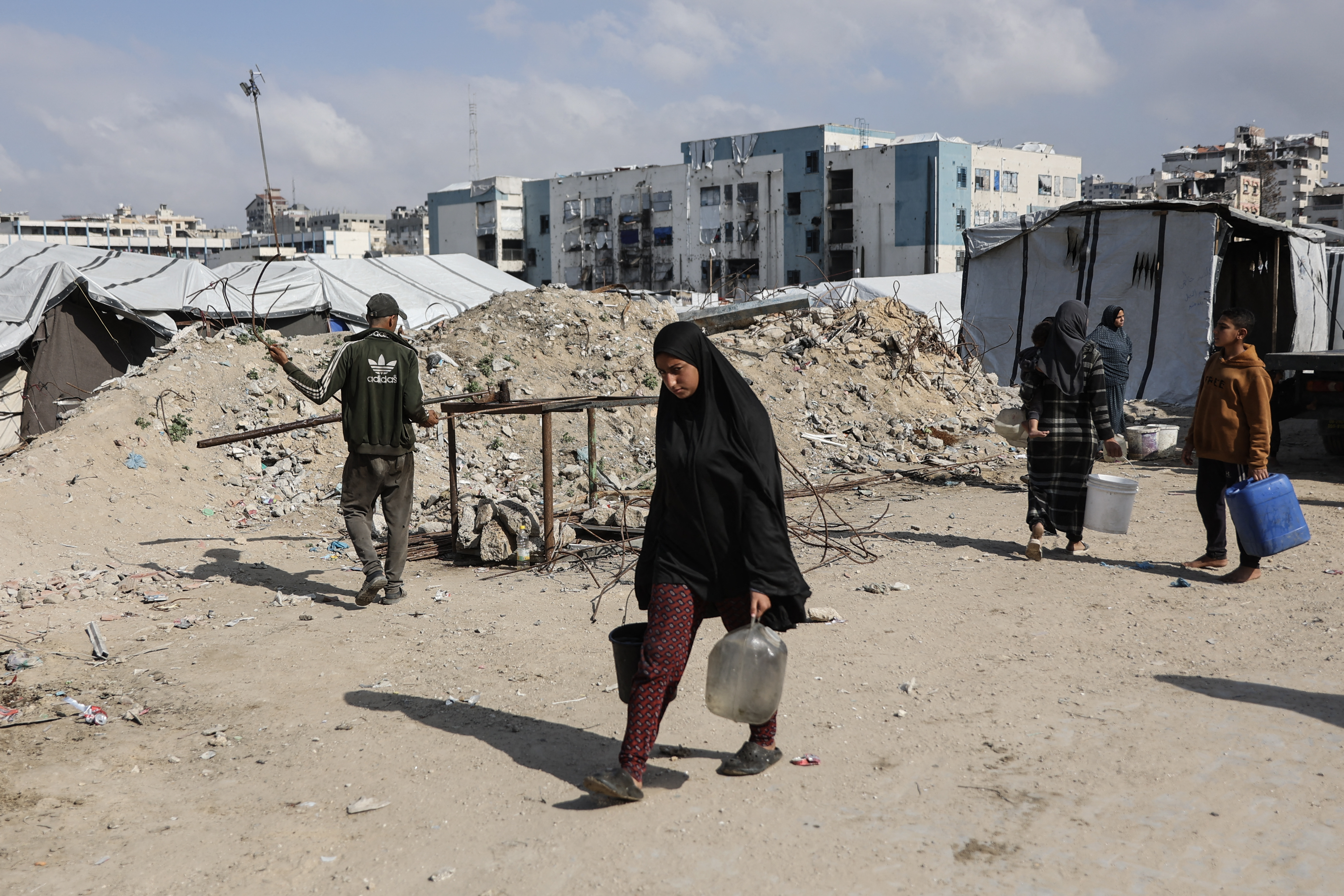 A displaced Palestinian woman carries water containers after filling them from mobile cisterns in the Al-Rimal shelter camp in Gaza City on February 21, 2026. Nearly all of Gaza's 2.2 million residents were displaced at least once during the more than two years of war between Israel and Hamas, sparked by the latter's unprecedented October 7 attack on Israel. (Photo by Omar AL-QATTAA / AFP)