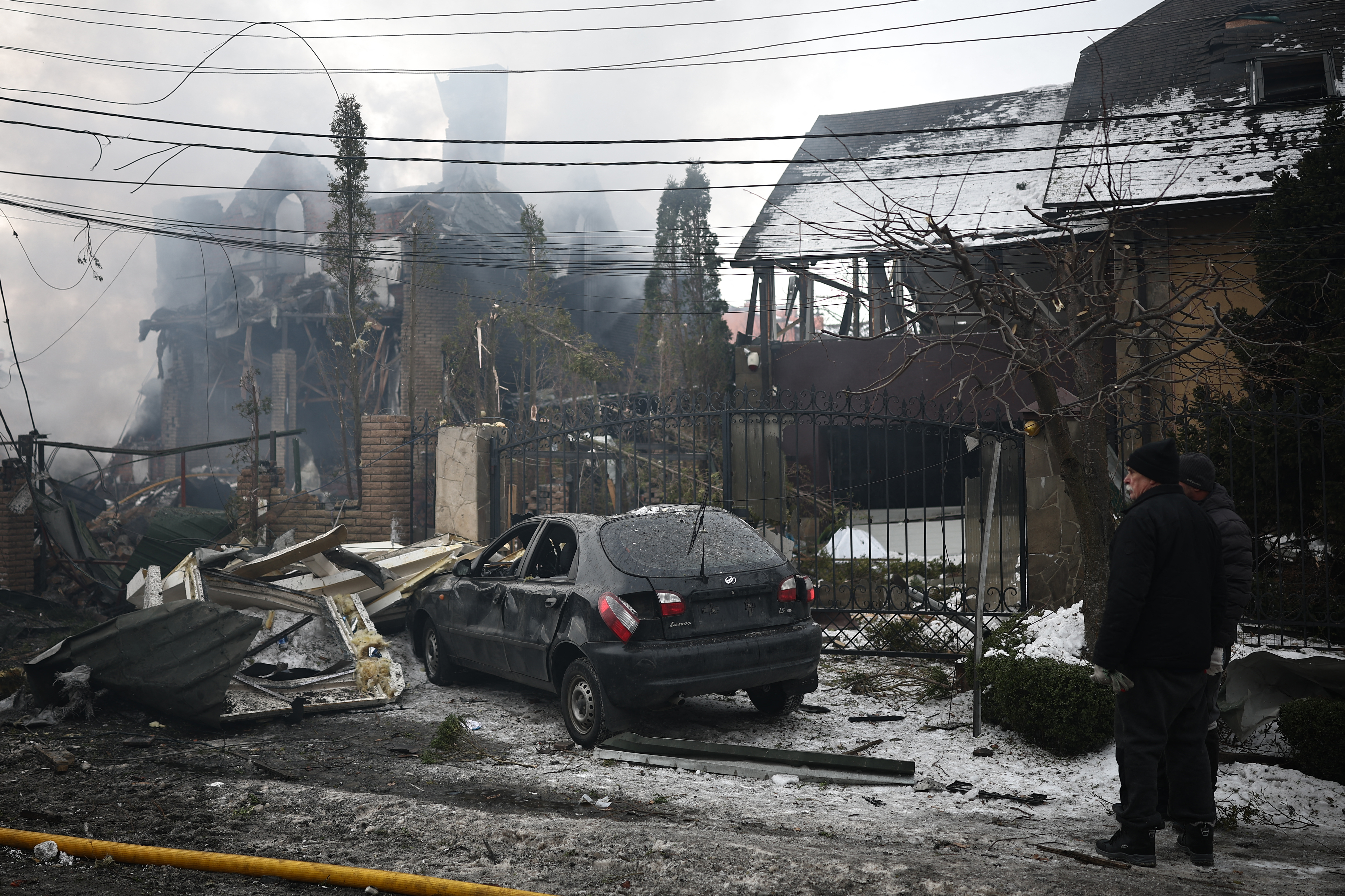 Local residents stand next to a damaged house and car at the site of an air attack in Sofiivska Borshchagivka, Kyiv region on February 22, 2026, amid the Russian invasion of Ukraine. Explosions rocked Ukraine's capital Kyiv with officials warning of a ballistic missile attack, just two days before the fourth anniversary of Russia's invasion. (Photo by Henry Nicholls / AFP)