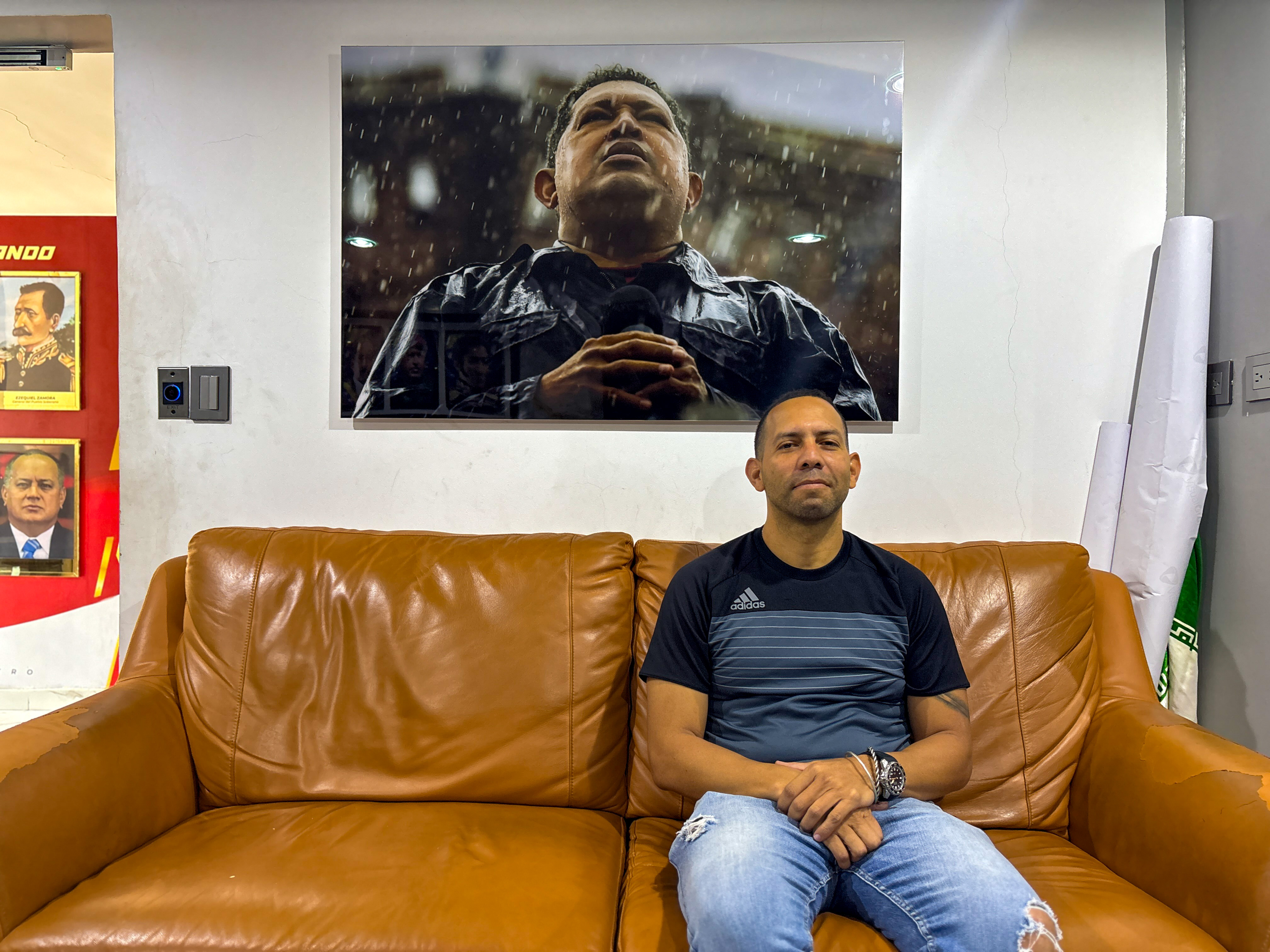 Jonsy Serrano sits on a couch below a portrait of Hugo Chavez