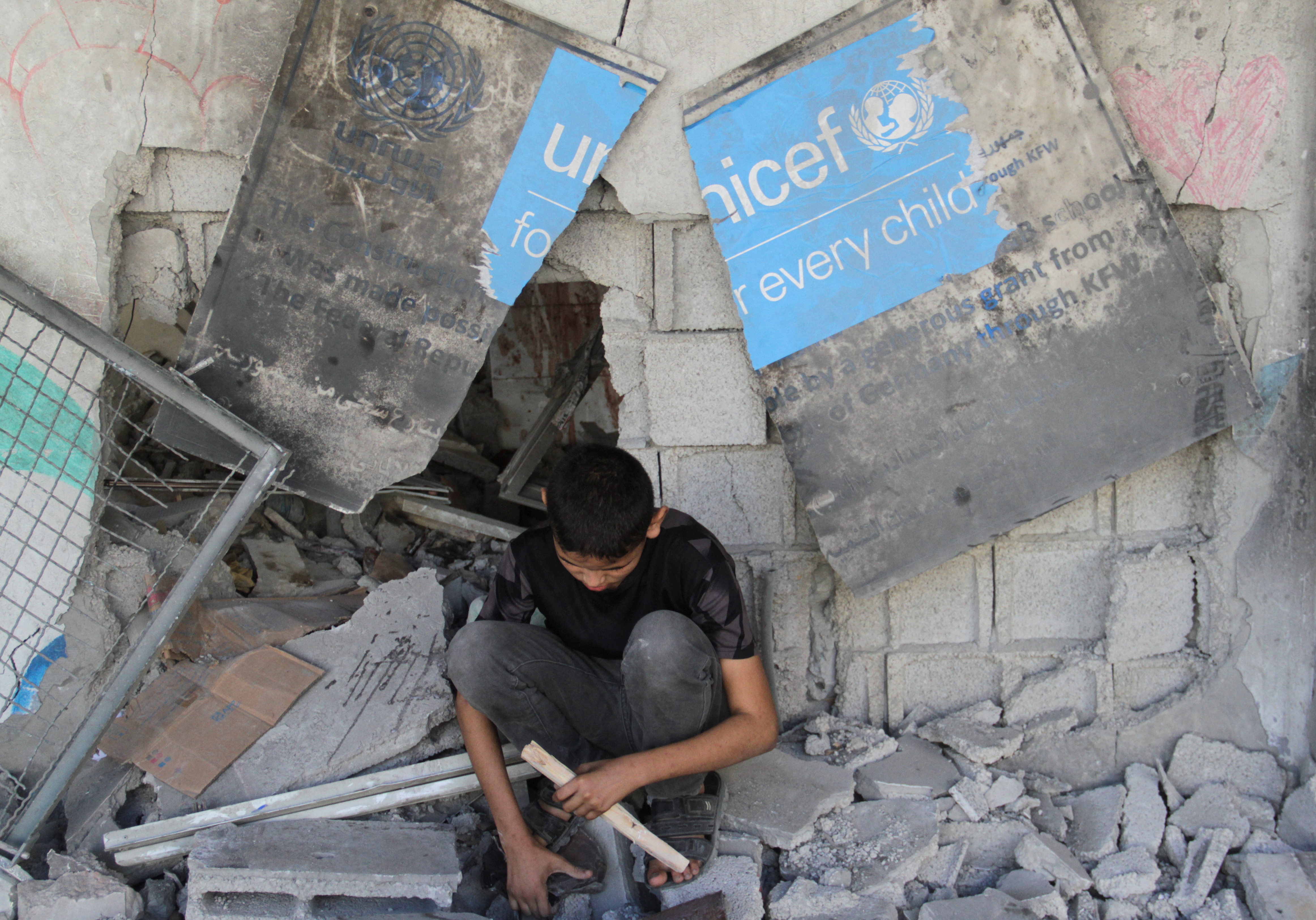 A boy sits under a broken UNICEF sign as Palestinians inspect the site of an Israeli strike on a school sheltering displaced people [File: Mahmoud Issa/Reuters]