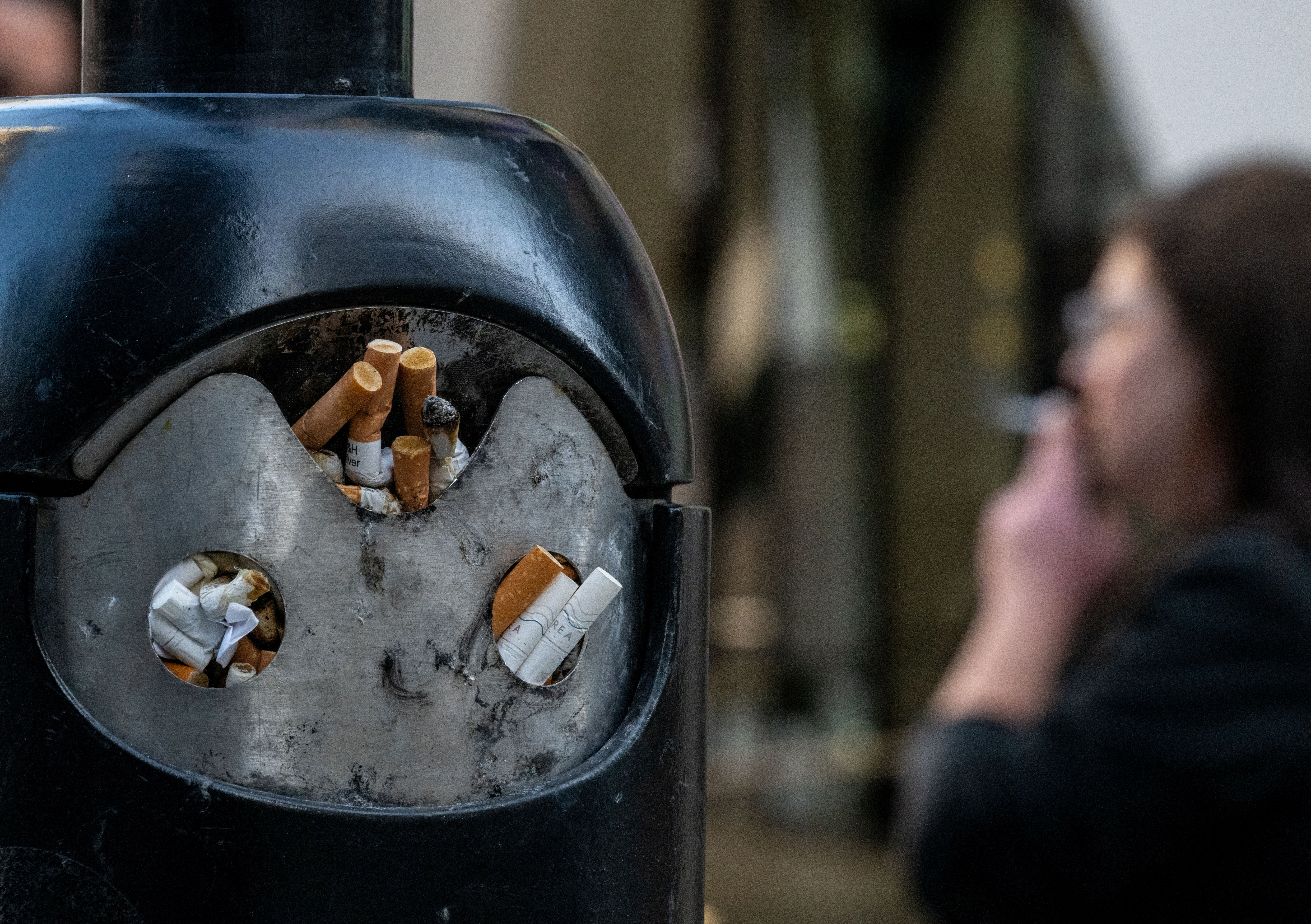 A smoker stands next to a bin for cigarette butts.