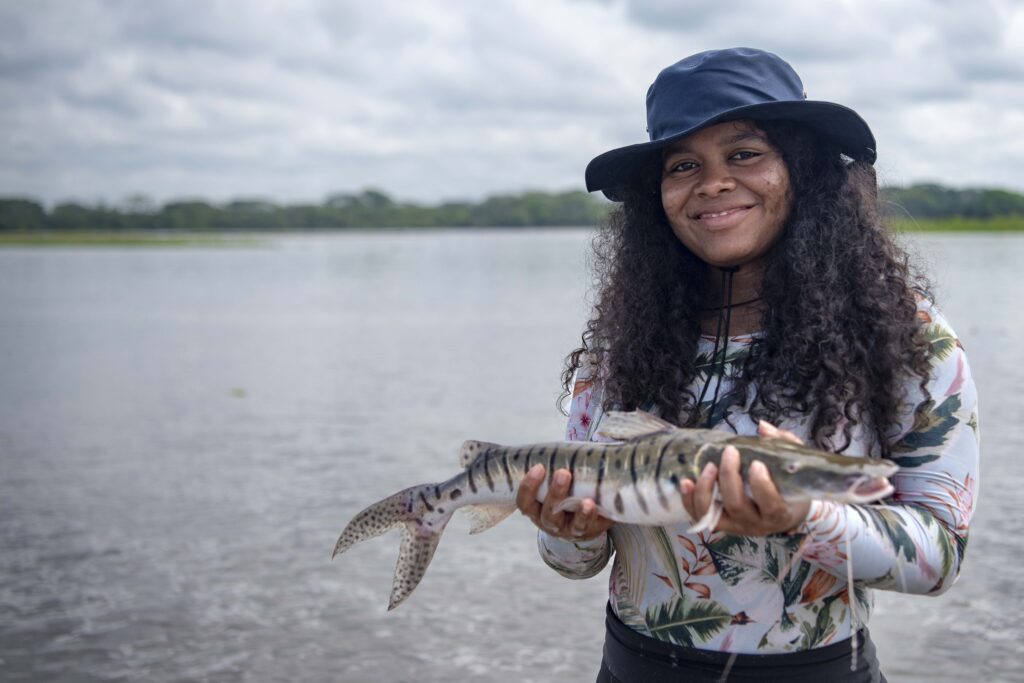 A young woman wearing a broad hat holds a fish next to a river, smiling 