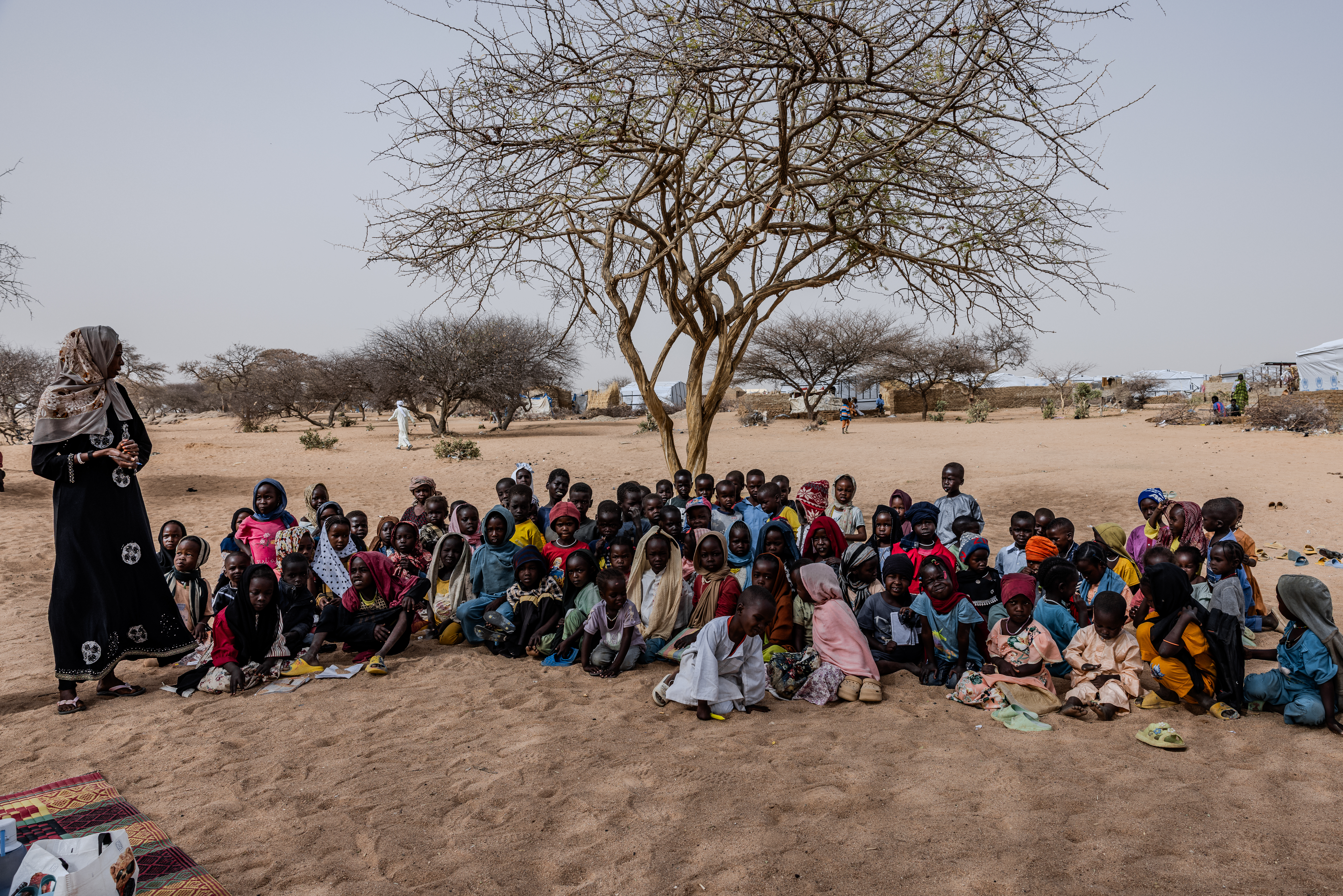 Crowd of children sits under a tree and listens to a teacher.