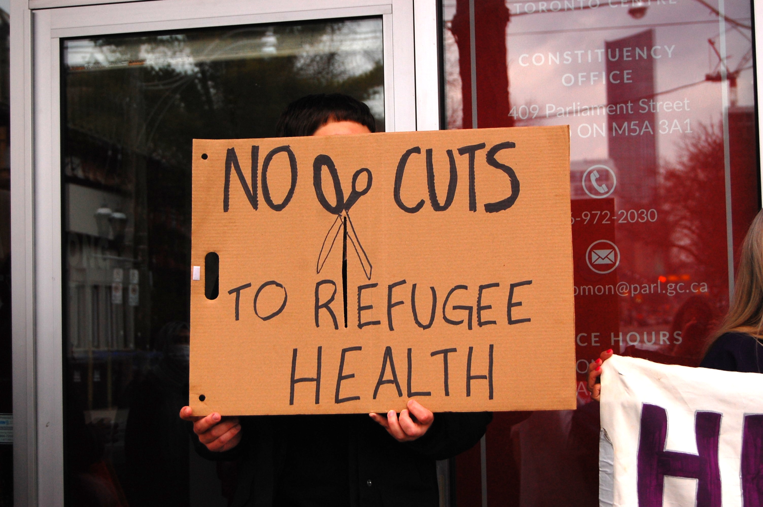 A protester holds a sign that reads, 'No cuts to refugee health'