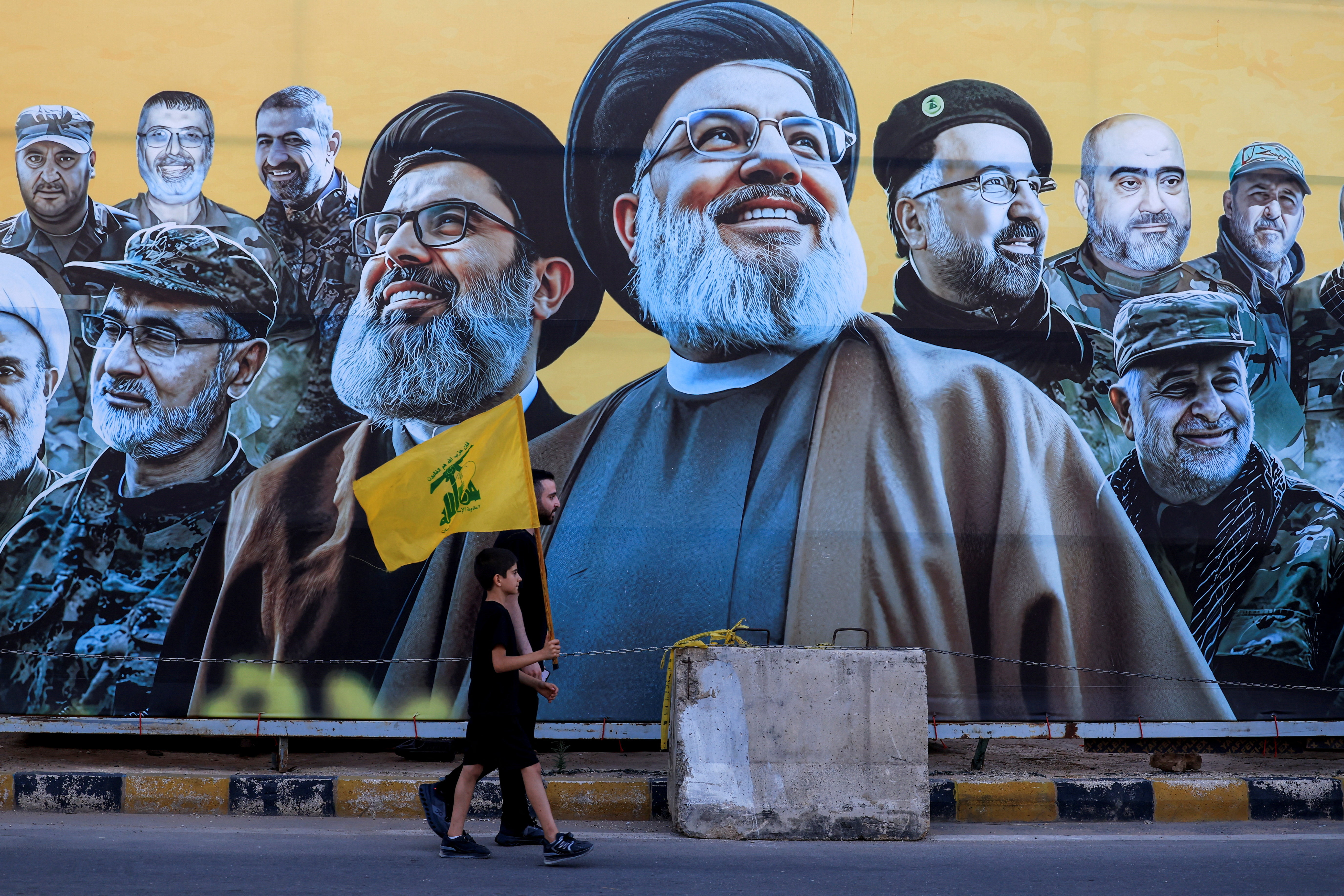 A man walks with a boy, carrying a Hezbollah flag, past a mural depicting former Hezbollah leader Hassan Nasrallah and other senior figures, on the day Hezbollah chief Naim Qassem delivers a televised speech, near the burial site of Hassan Nasrallah on the outskirts of Beirut, Lebanon, June 17, 2026.