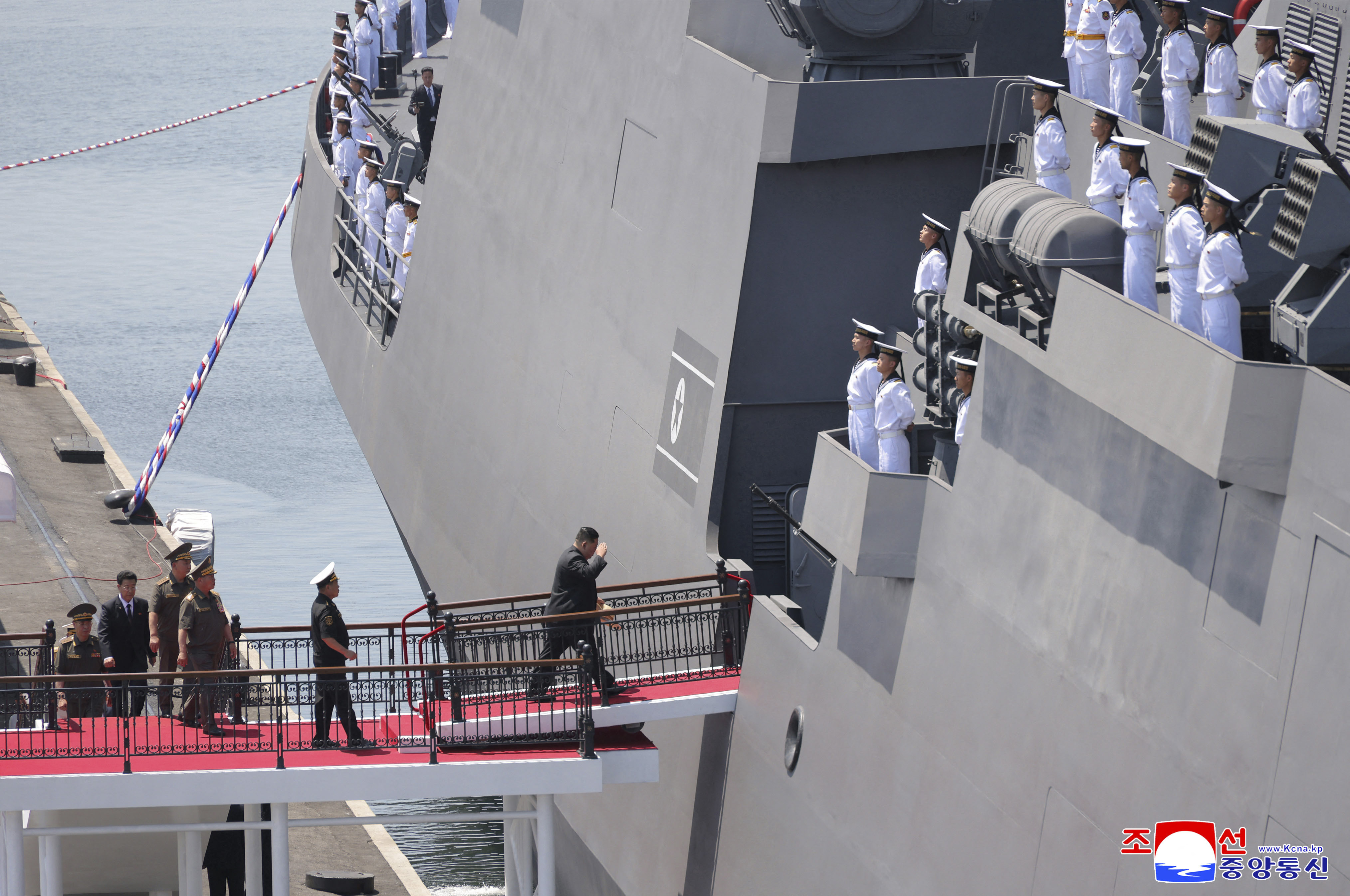 Figures walk across a red-carpeted gangway onto a large grey warship as rows of uniformed crew stand on the deck above.