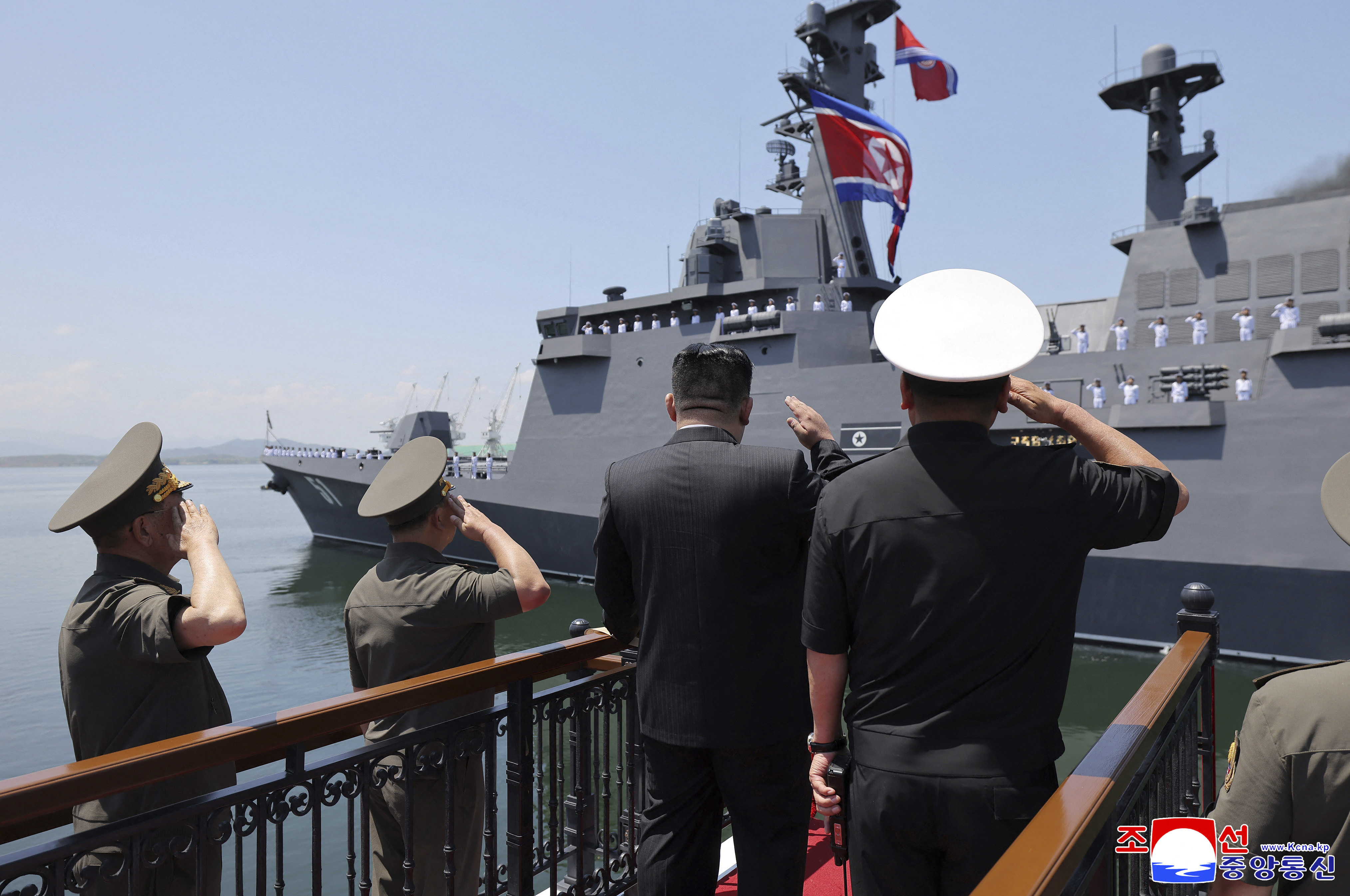 Kim Jong Un raises his hand in salute toward a large grey warship at a naval ceremony, with people standing on either side of him.