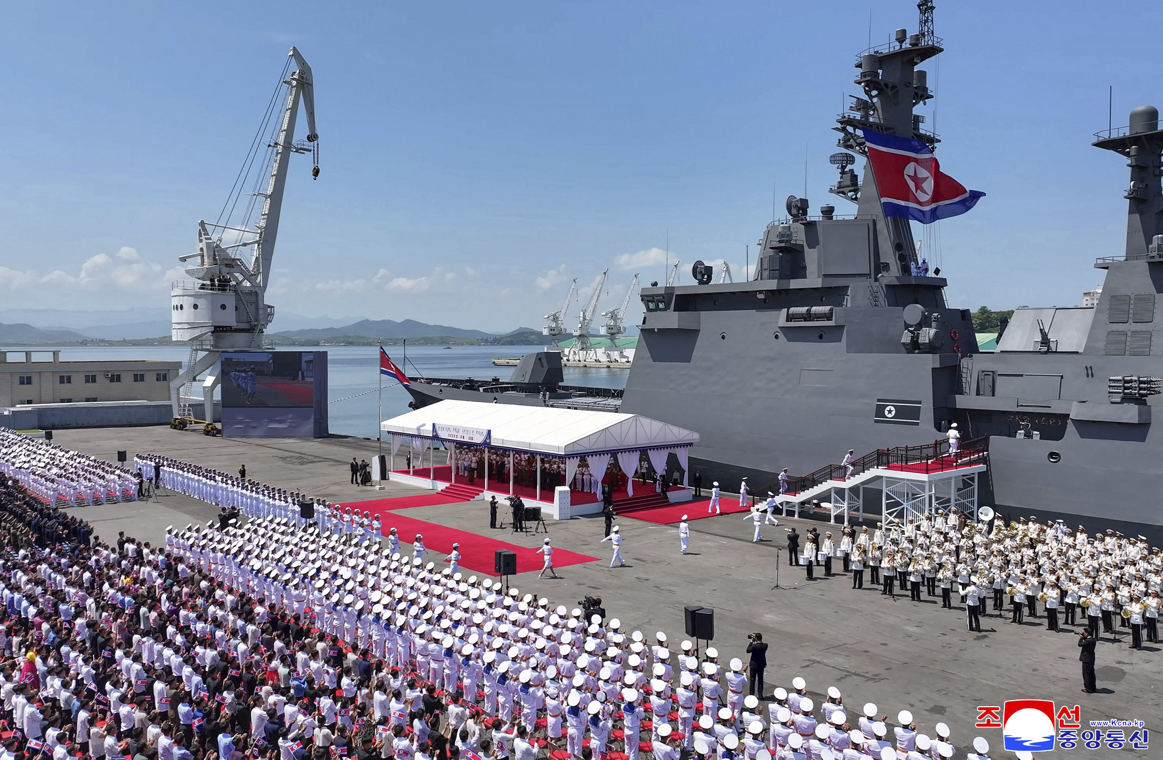 North Korean leader Kim Jong Un stands at a podium addressing a naval commissioning ceremony, with a large grey warship behind him.