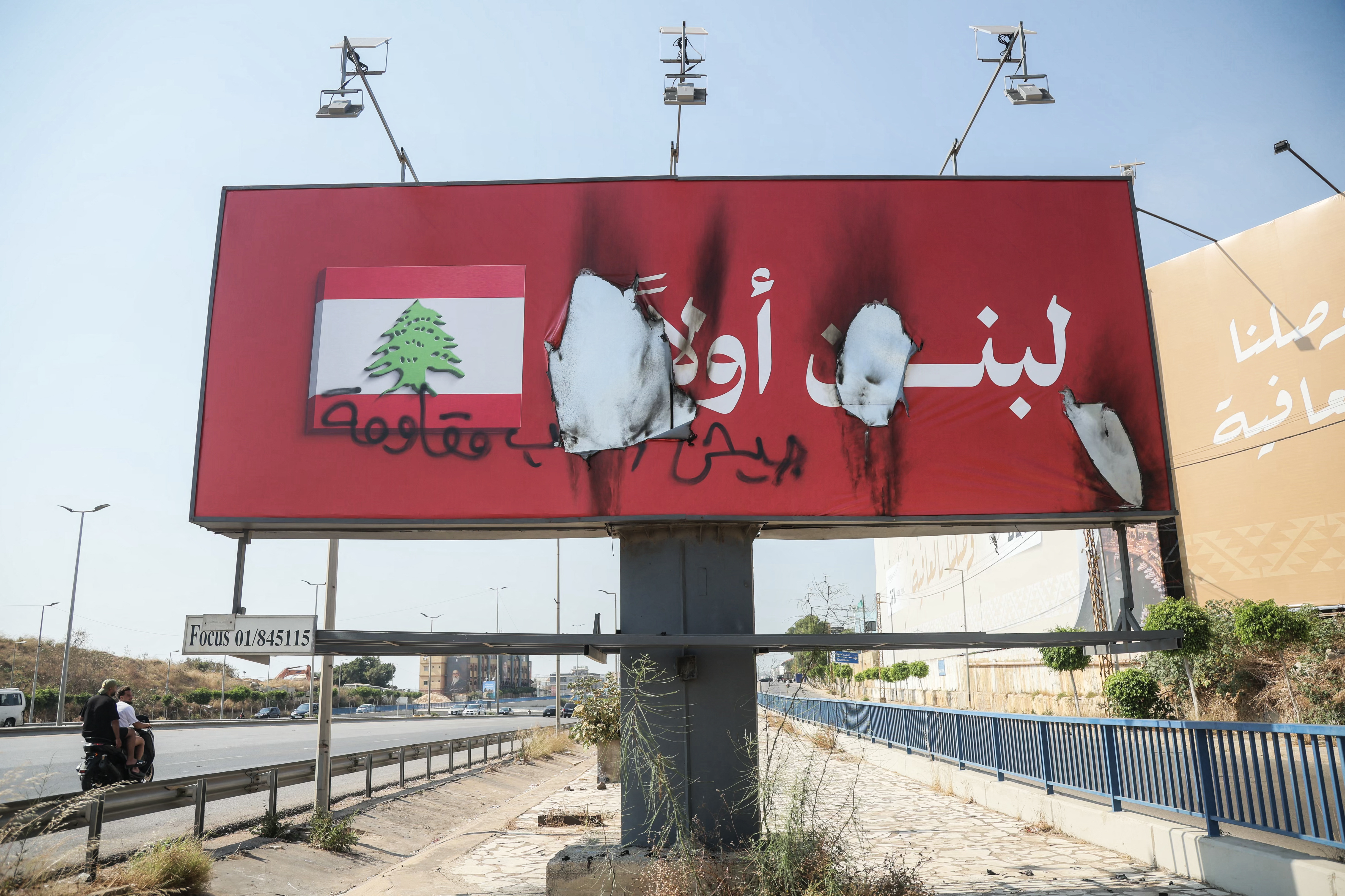 Vehicles drive past a billboard that reads in Arabic "Lebanon first" (in white) after it was defaced with the words "Army, People, Resistance" and set on fire along Beirut's airport road near Tehran-backed Hezbollah's stronghold, amidst tensions following Lebanon's framework agreement with Israel on June 28, 2026. The current billboards recently replaced previous billboards that read Thank you, loyal Iran, Hezbollah's main backer. The militant group has rejected the agreement, which was signed in Washington on June 26, after five rounds of talks and aims to pave the way to peace between the neighbours, includes plans to disarm Hezbollah. (Photo by ibrahim AMRO / AFP) /