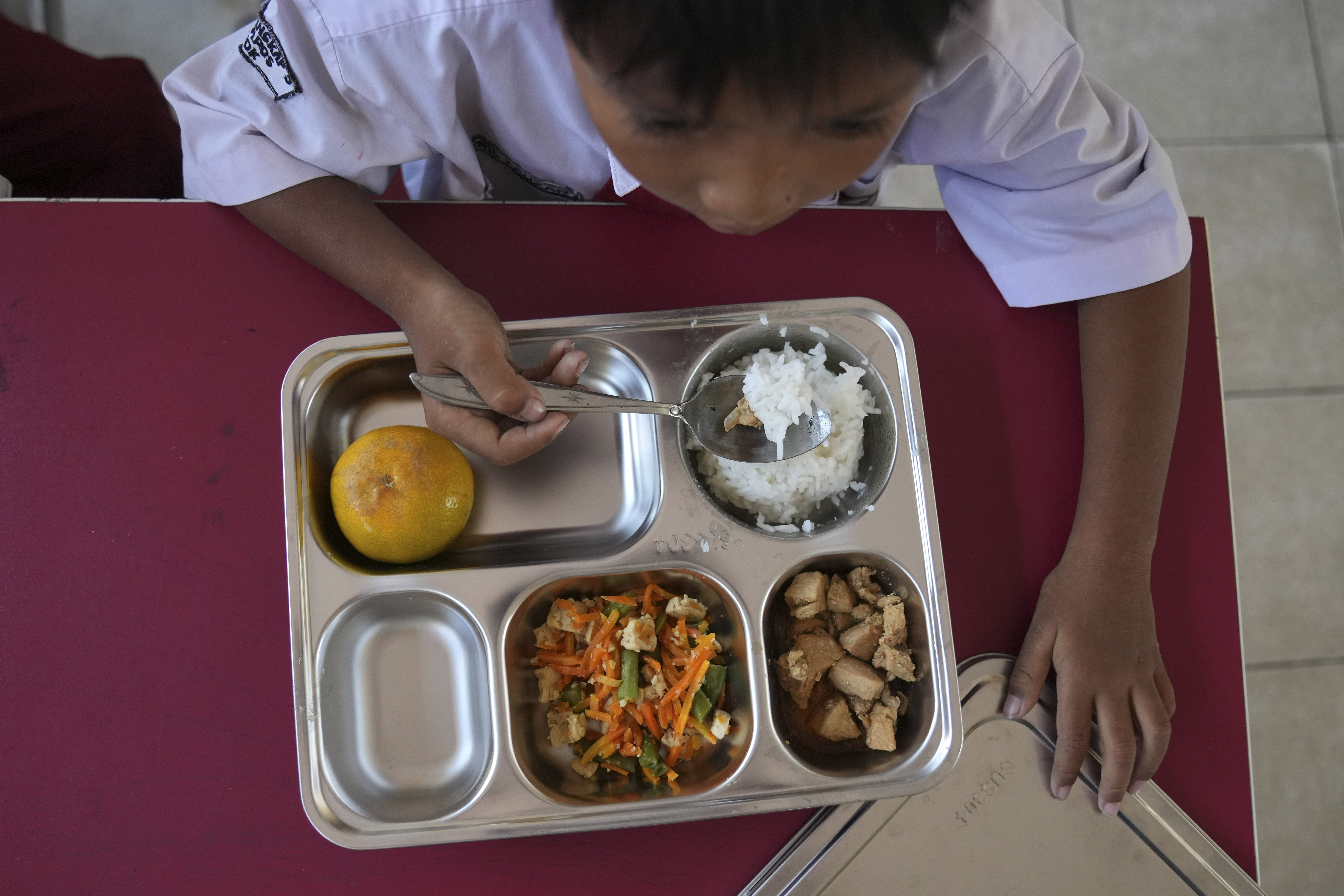 A young student in school uniform eats from a meal tray at a classroom desk, with other children eating around him