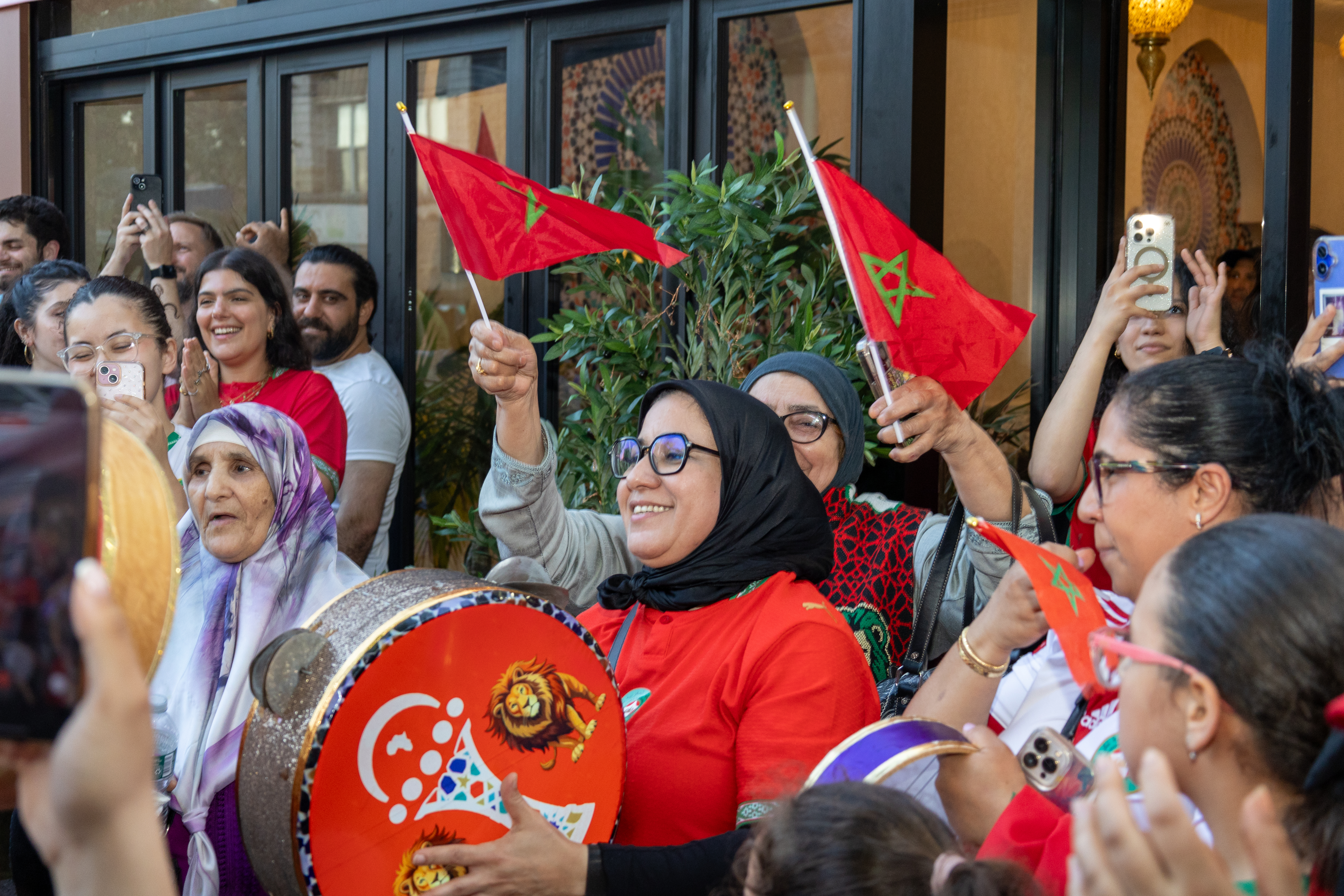 A crowd watches a football match on an outdoor screen, some holding red Moroccan flags.