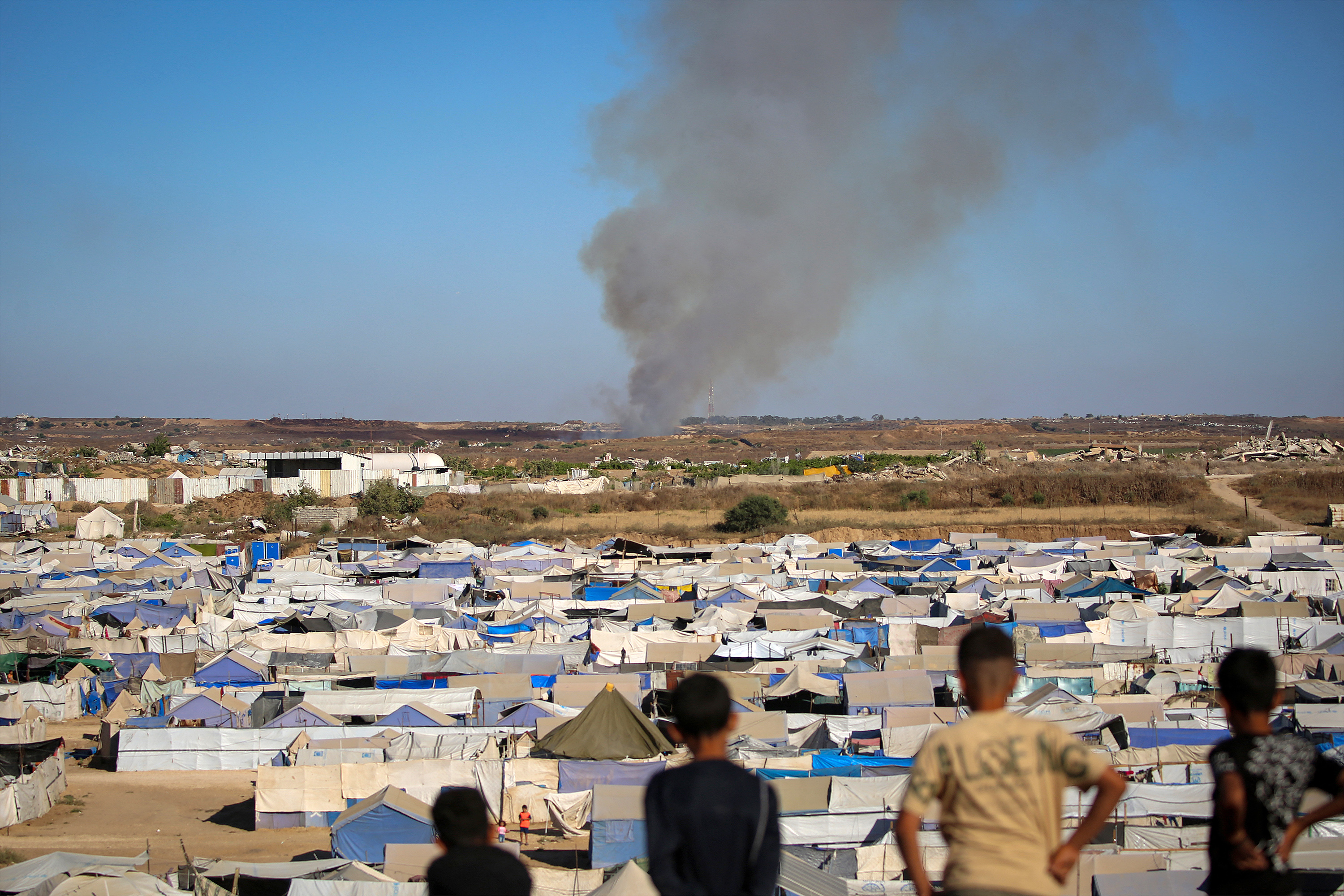Children watch from atop a promontory overlooking shelters for people displaced by war, as smoke billows in the background.