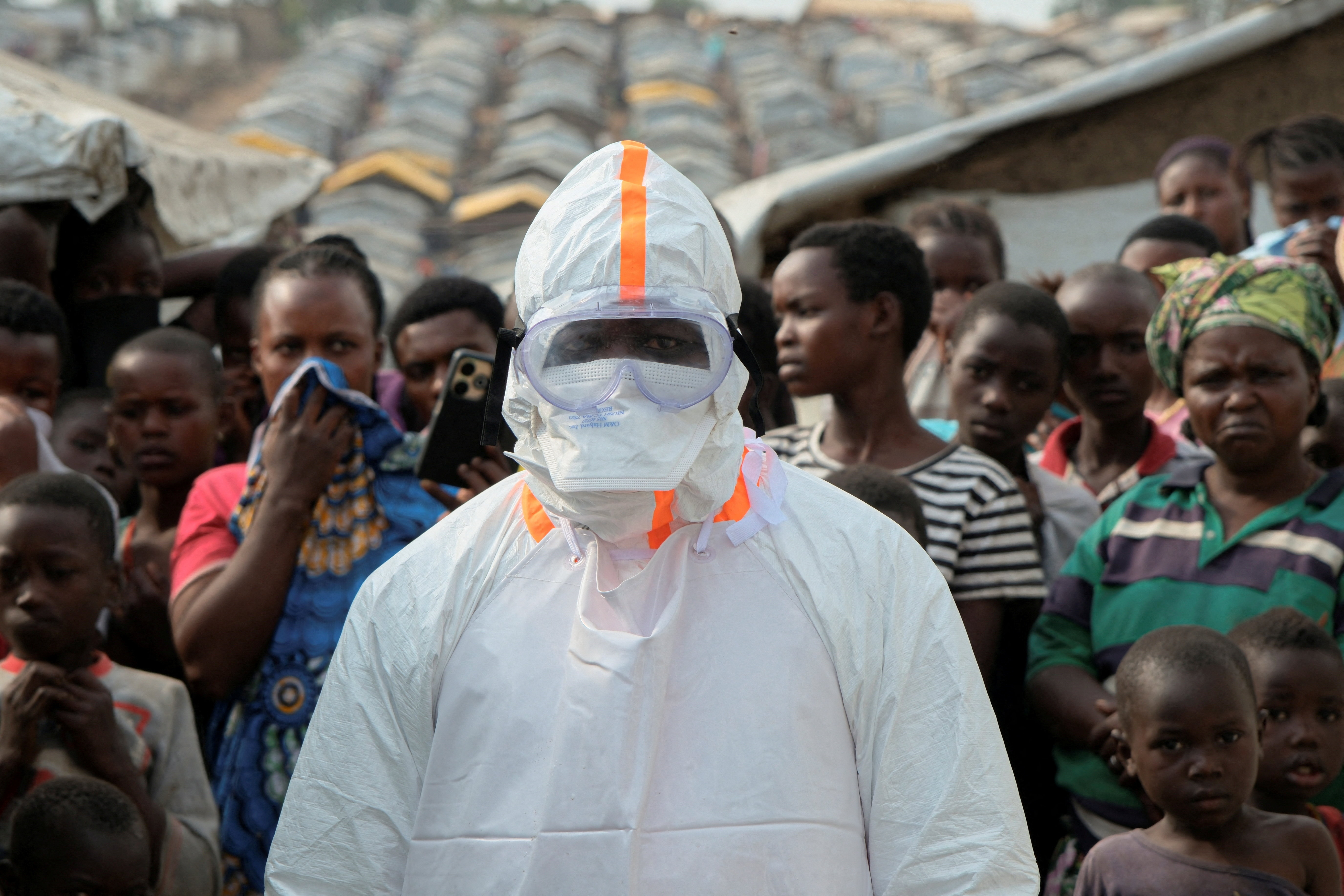 A health worker in full protective gear stands near a group of people gathered for a burial at a displaced persons camp.