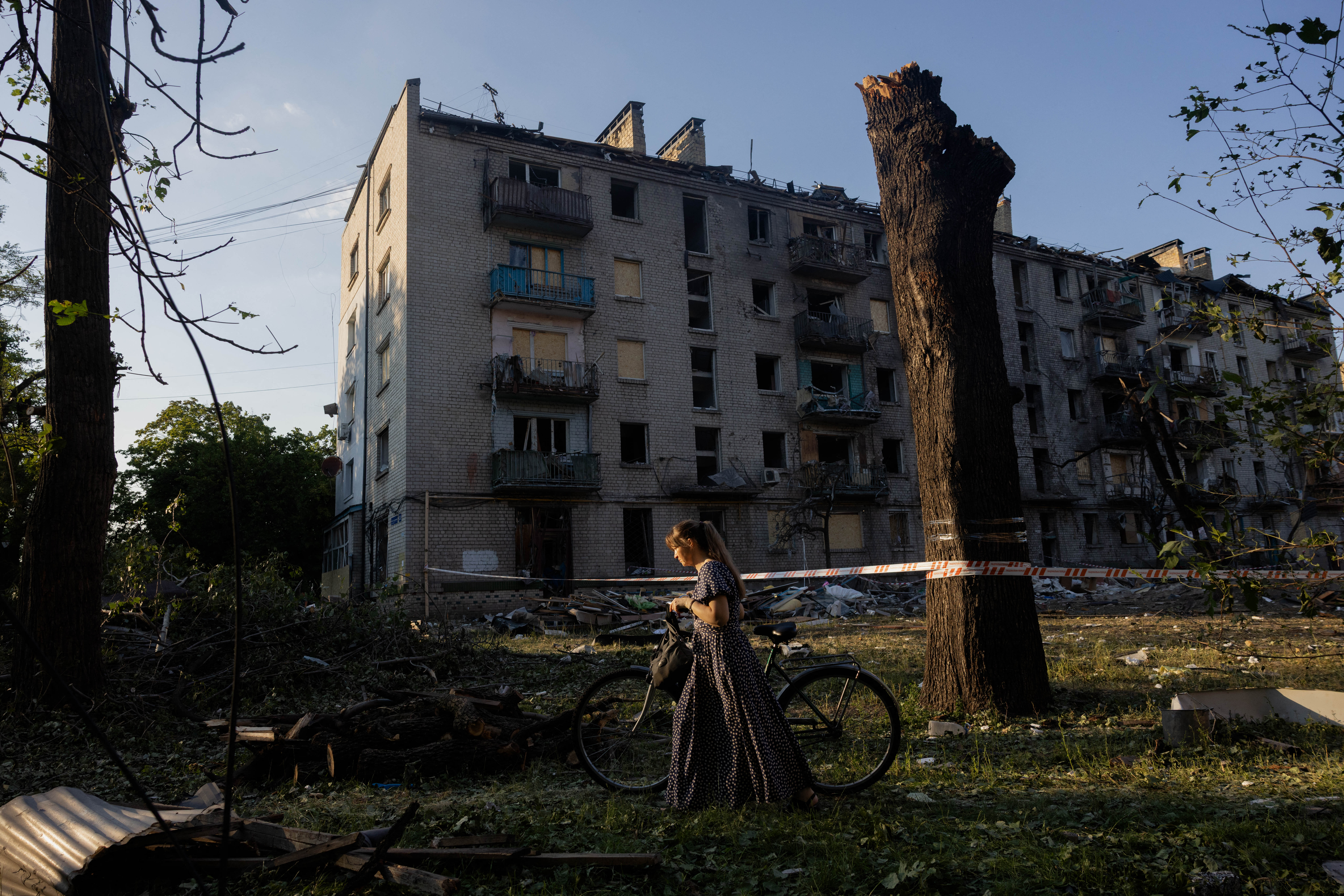 A woman with a bicycle walks by a damaged building.