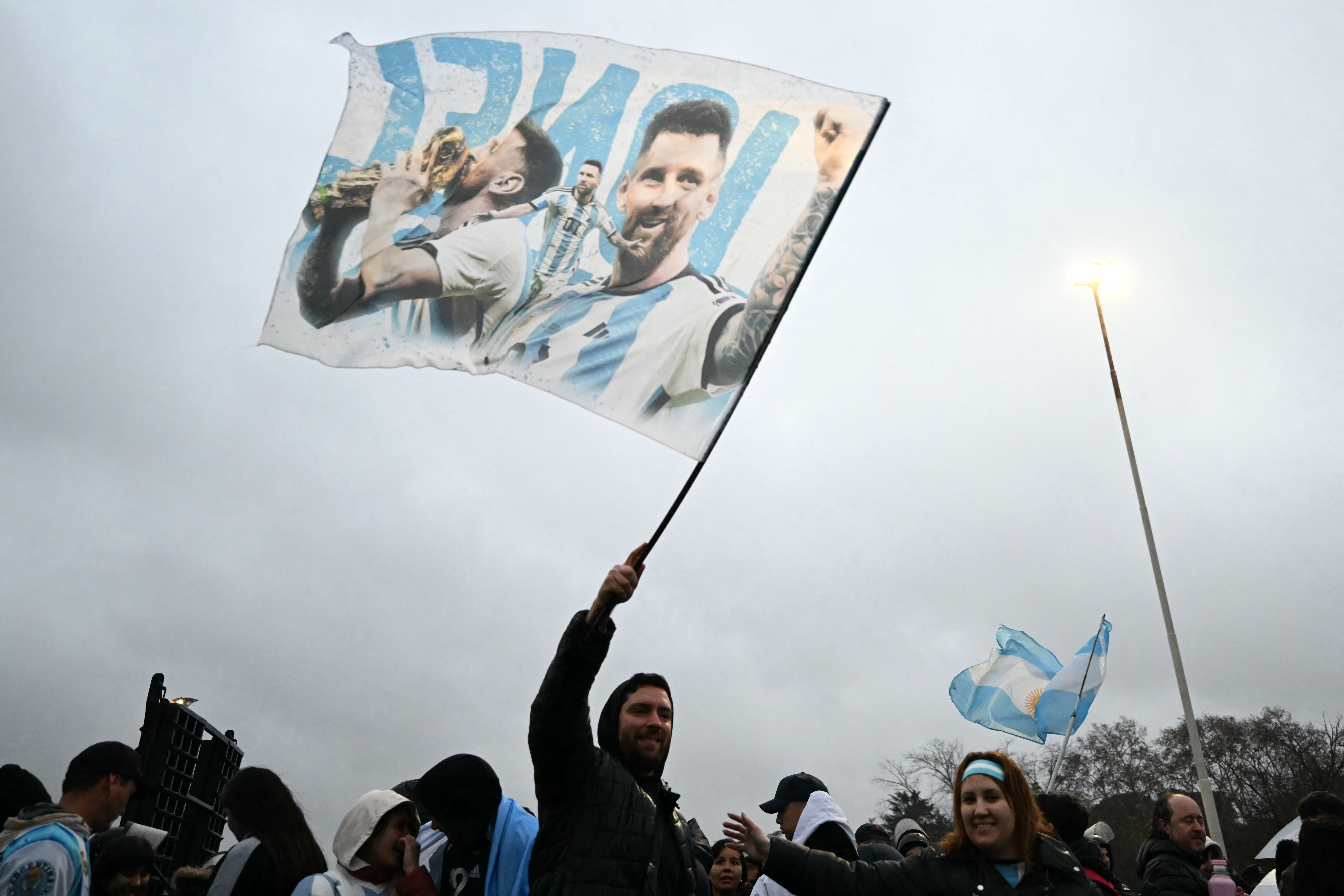 A fan waves a flag bearing images of Argentina's forward Lionel Messi near the Argentine Football Association (AFA) training camp in Ezeiza, Buenos Aires Province, on July 20, 2026, while waiting for the arrival of Argentina's national football team from the United States following its defeat in the 2026 FIFA World Cup final. (Photo by Luis ROBAYO / AFP)