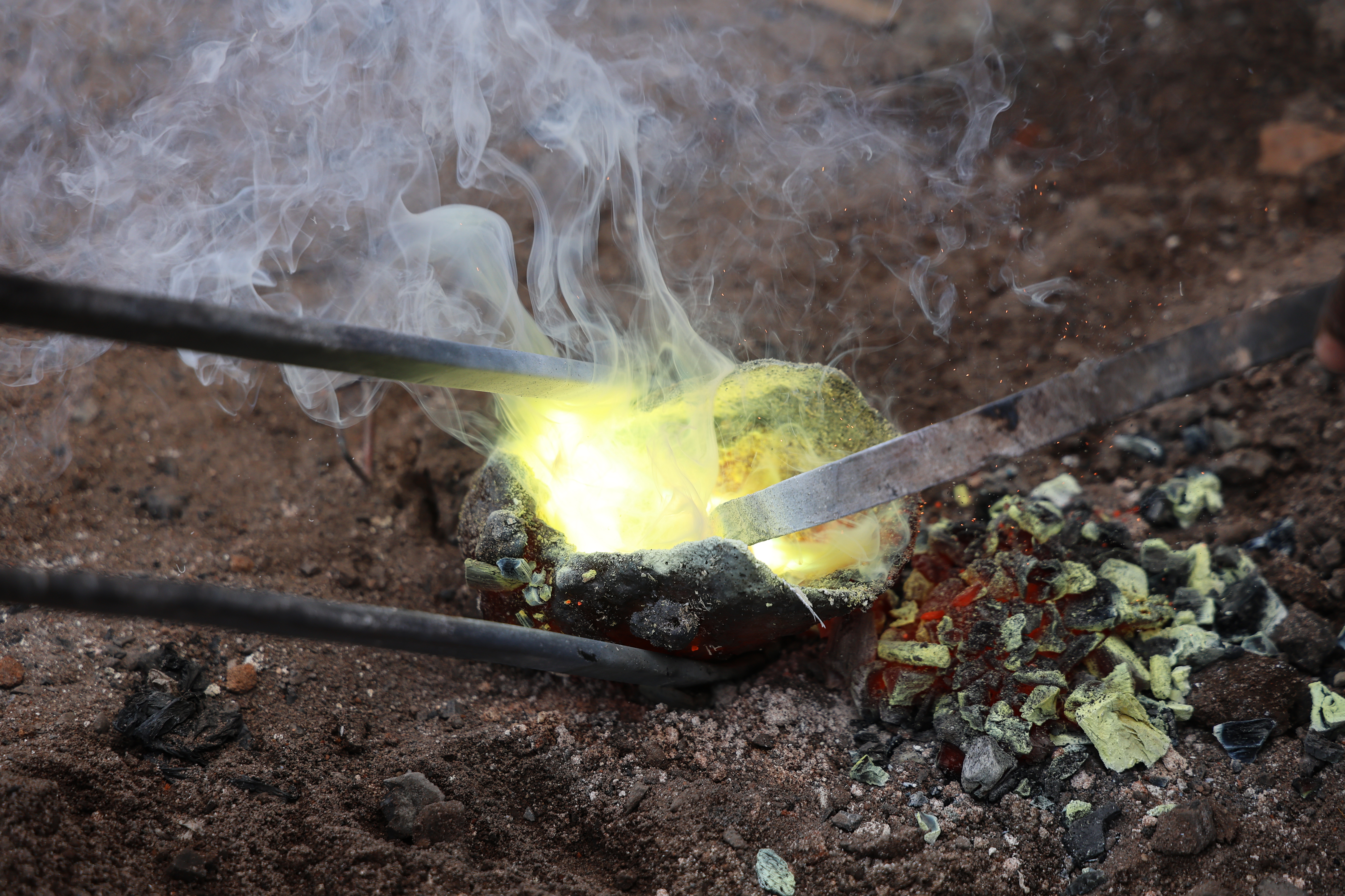A crucible of molten bronze rests above charcoal embers before artisans pour the metal into clay moulds using long iron tongs.