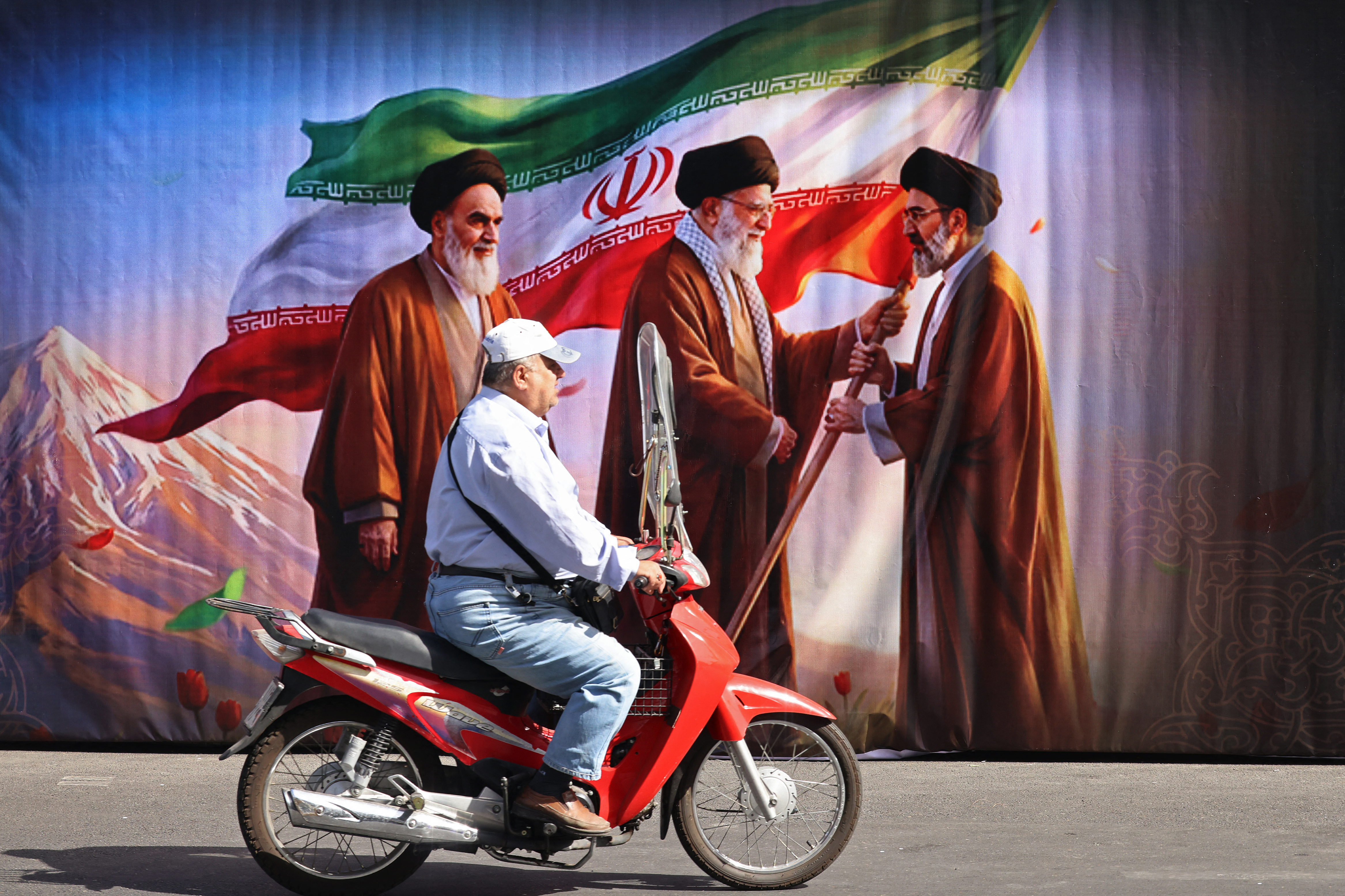 A man rides a scooter past a billboard featuring the images of the late founder of the Islamic Revolution supreme leader Ayatollah Ruhollah Khomeini (L), the Iran's slain supreme leader Ali Khamenei (C) and his son, the current supreme leader Mojtaba Khamenei, along the street in Tehran on July 3, 2026. Preparations for the funeral of Iran's slain supreme leader Ali Khamenei were in full swing on July 3, with authorities expecting millions and a coterie of foreign dignitaries to attend the official ceremony. (Photo by ATTA KENARE / AFP) /