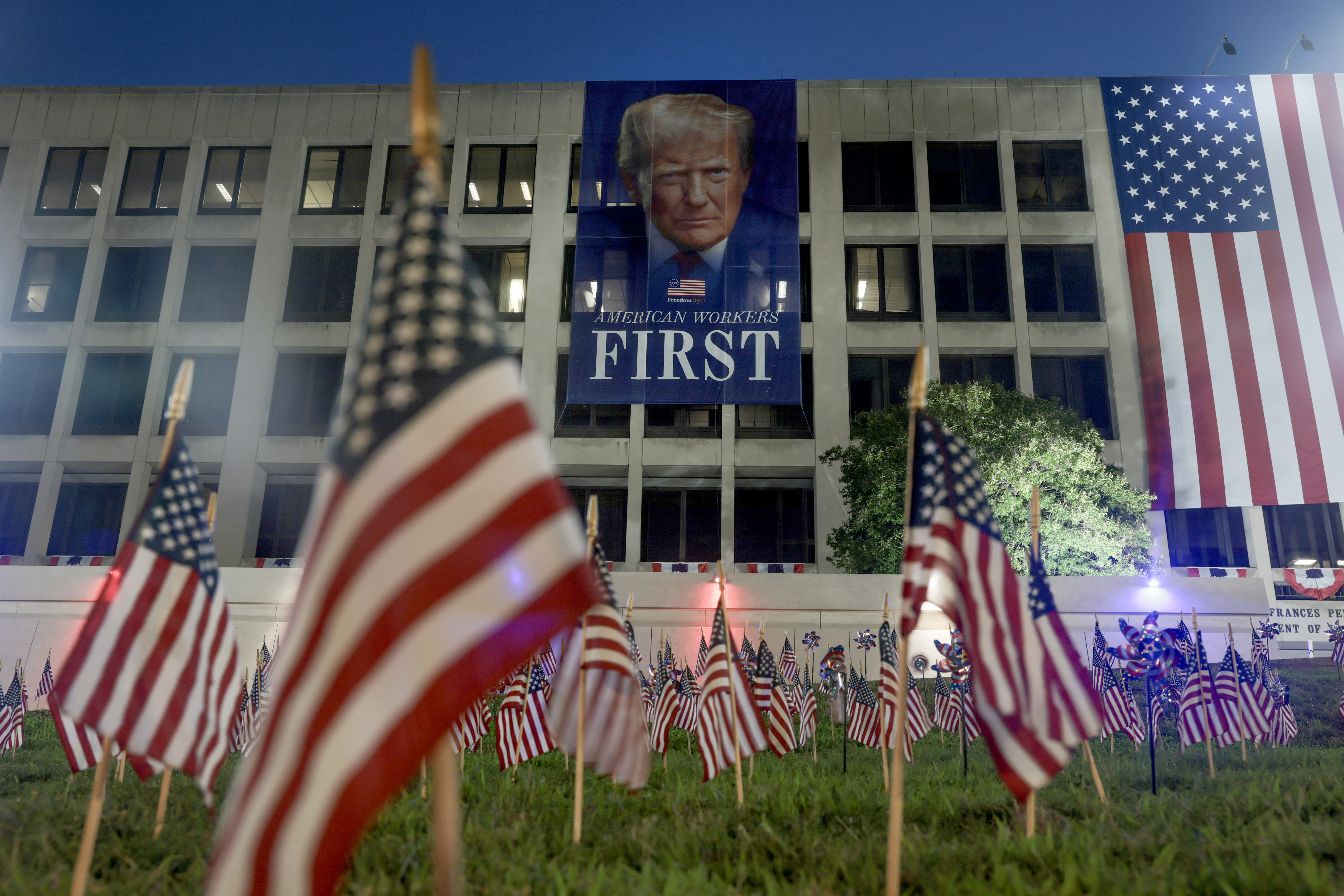 WASHINGTON, DC - JULY 03: American flags are planted in the ground in front of a banner of U.S. President Donald Trump, hanging from the U.S. Department of Labor building, ahead of July 4th festivities on July 03, 2026, in Washington, DC. A fireworks show will begin around 10:30 p.m on July 4th as the city deals with extreme heat warnings. Joe Raedle/Getty Images/AFP (Photo by JOE RAEDLE / GETTY IMAGES NORTH AMERICA / Getty Images via AFP)