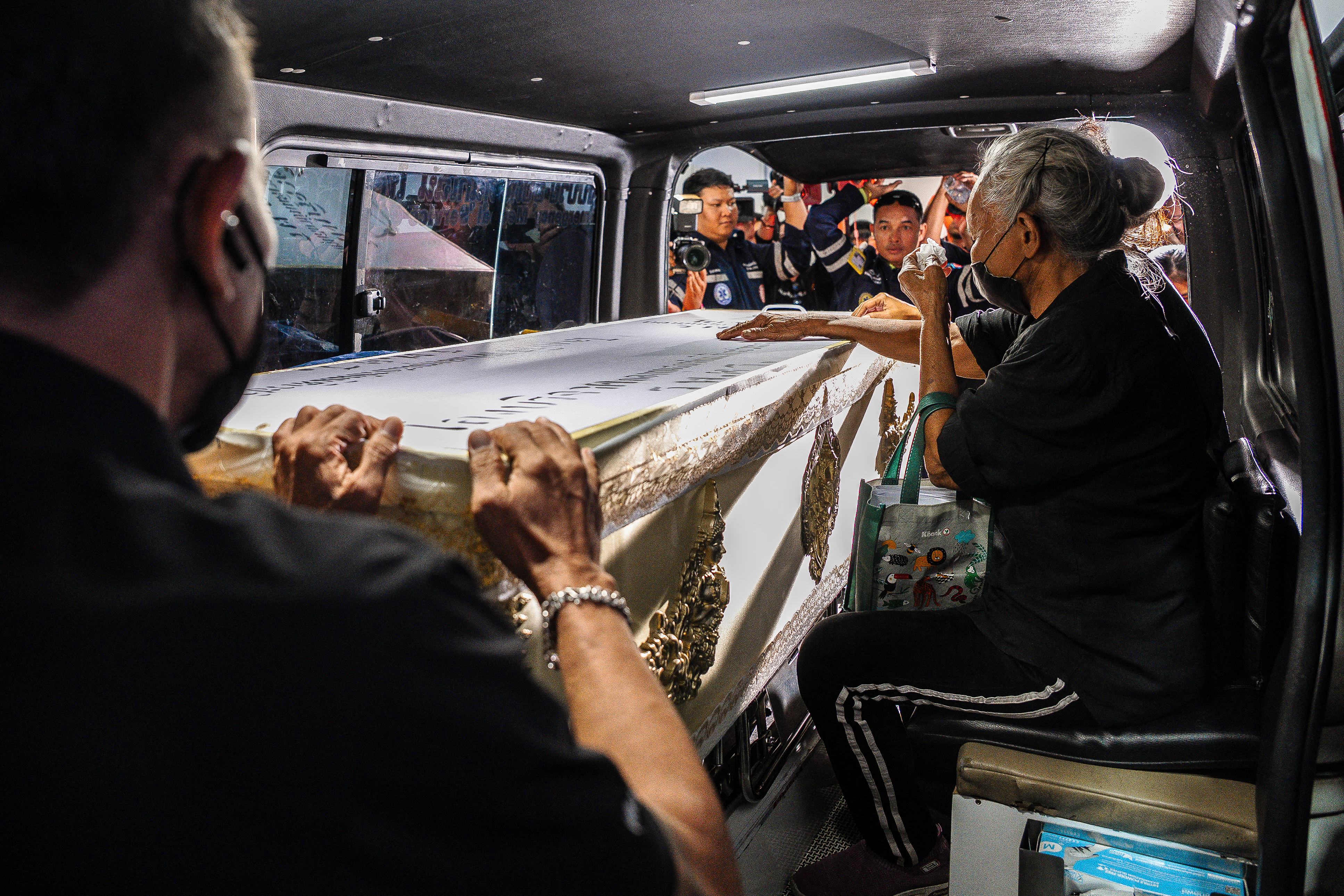 A person in mourning stands beside a coffin inside a hearse, with hospital staff nearby