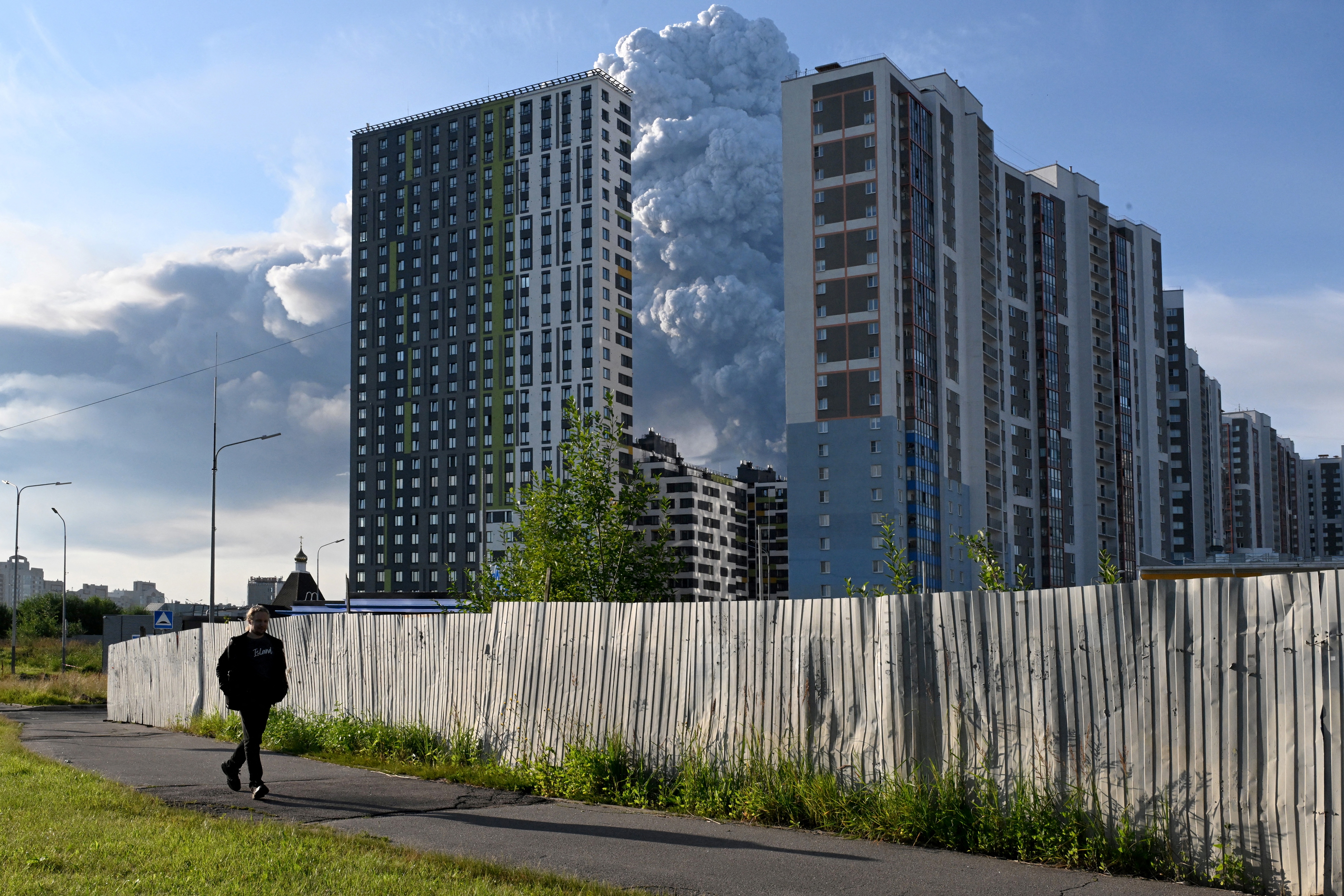 TOPSHOT - A man walks past residential buildings as smoke rises from a warehouse for a Russian e-commerce giant near Saint Petersburg after being hit by Ukrainian drones early on July 24, 2026 morning, triggering a fire that sent smoke pouring over the historic city, according to local officials.