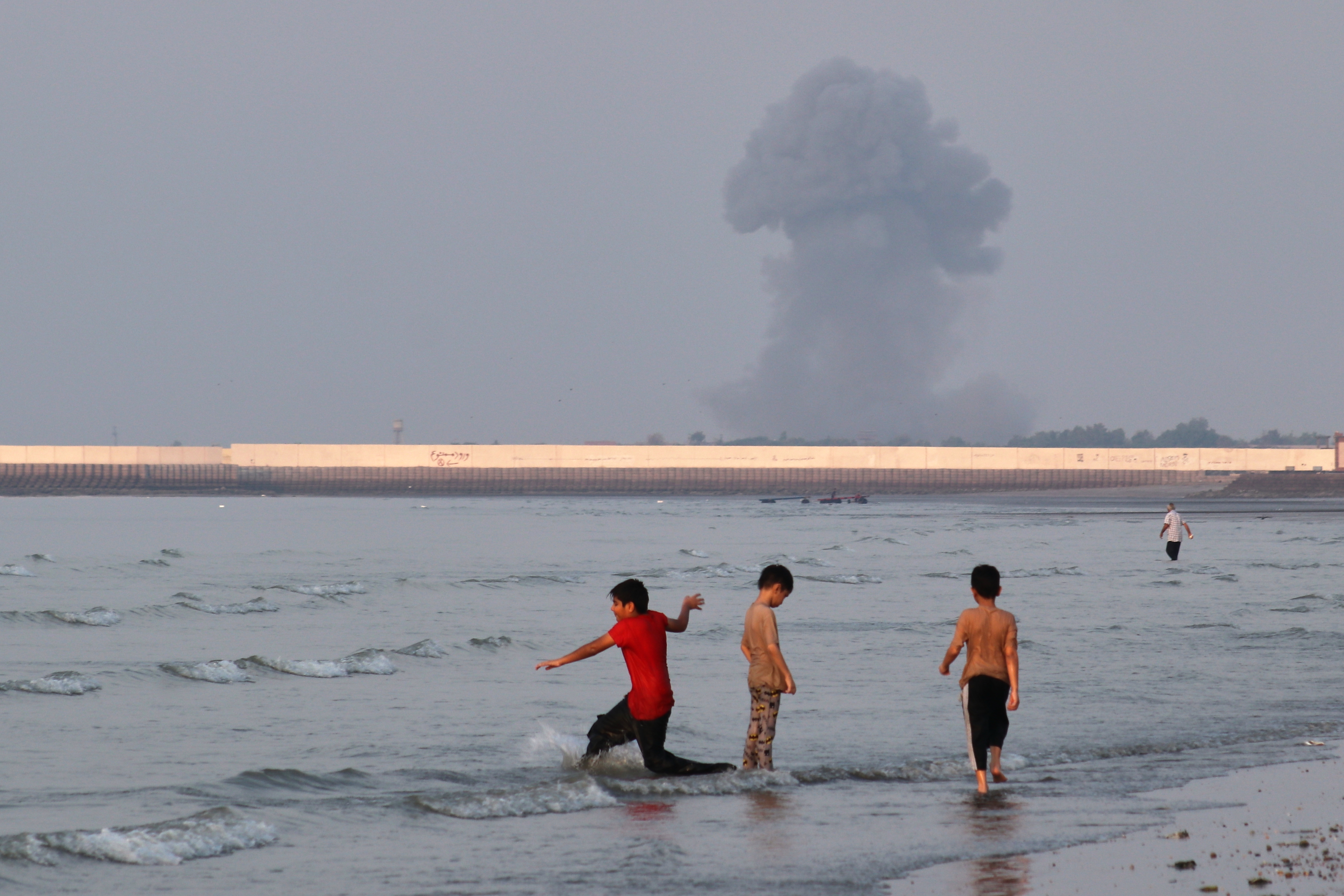 Three boys play in the shallow waters of the Strait of Hormuz, as a plume of smoke rises from an explosion in the background, off Bandar Abbas, Iran, Monday, July 13, 2026. (Razieh Poudat/ISNA via AP)
