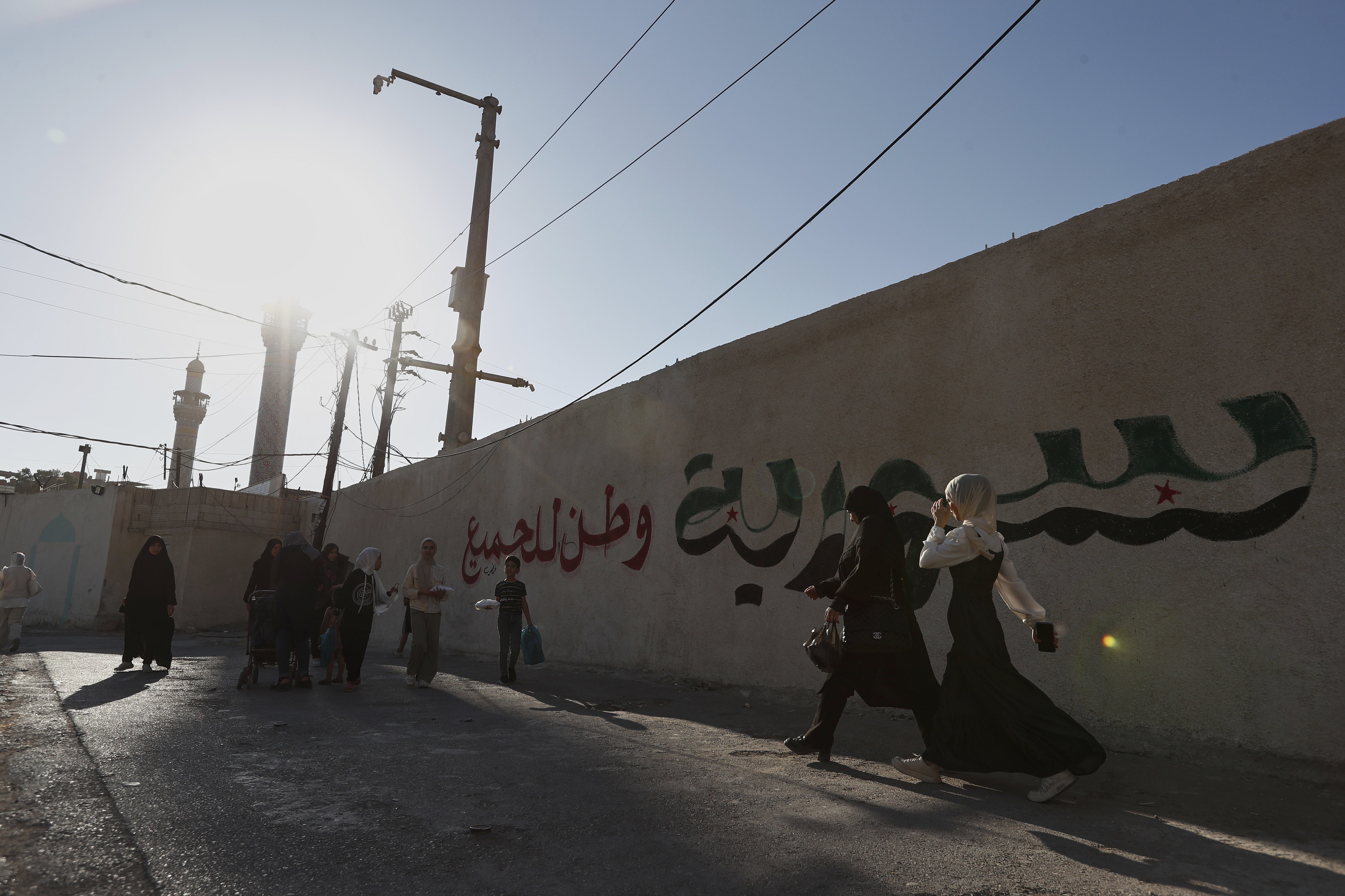 Syrian women pass next to a wall with Arabic words that read:'Syria is nation for all", outside the shrine of al-Sayydah Zeinab, the grand daughter of Prophet Mohammed, where Shiite worshippers attend Ashoura ritual, south of Damascus, Syria, Thursday, July 3, 2025. (AP Photo/Omar Sanadiki)