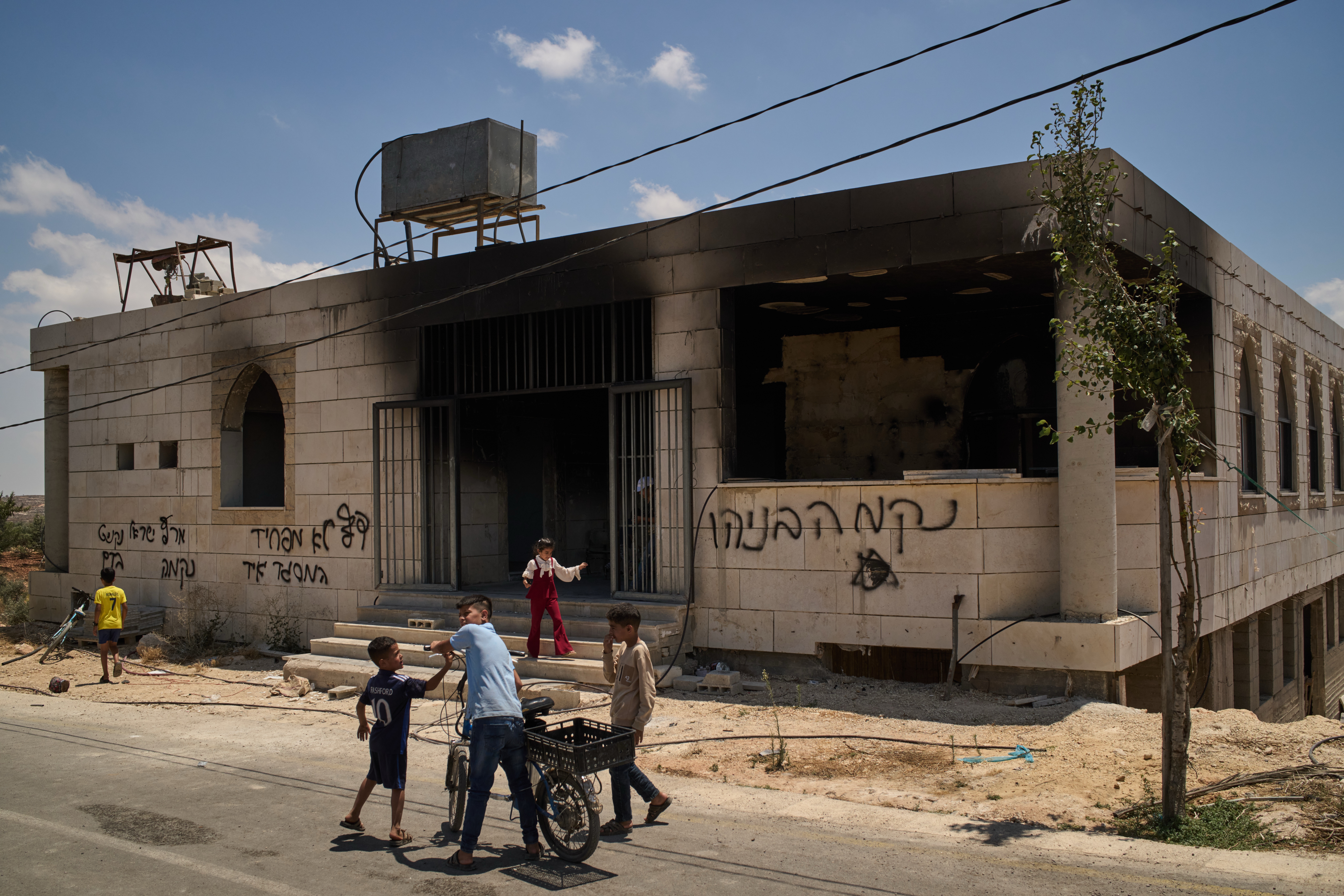 Children stand outside a mosque that was burned during an attack by Israeli settlers early in the morning in the West Bank village of Qusra, Sunday, July 26, 2026, as on its wall a Hebrew graffiti reads "Revenge Benayahu", referring to an Israeli settler killed during clashes with Palestinians in the village of Tell, last Friday. (AP Photo/Leo Correa)