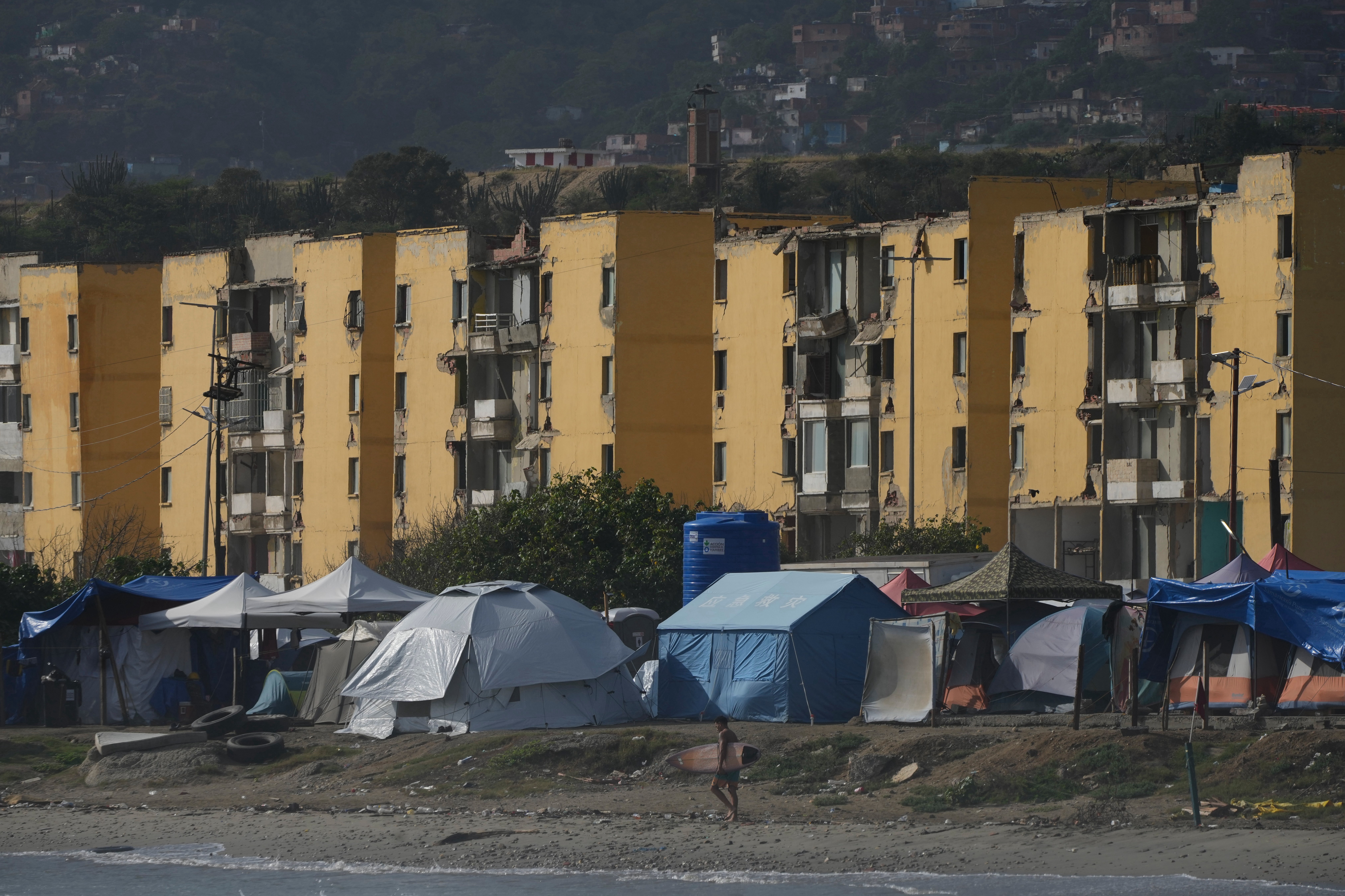 A surfer walks past a tent encampment on his way to a traditional "Paddle Out" ceremony to pay tribute to earthquake victims, in La Guaira, Venezuela, Saturday, Aug. 1, 2026. (AP Photo/Ariana Cubillos)
