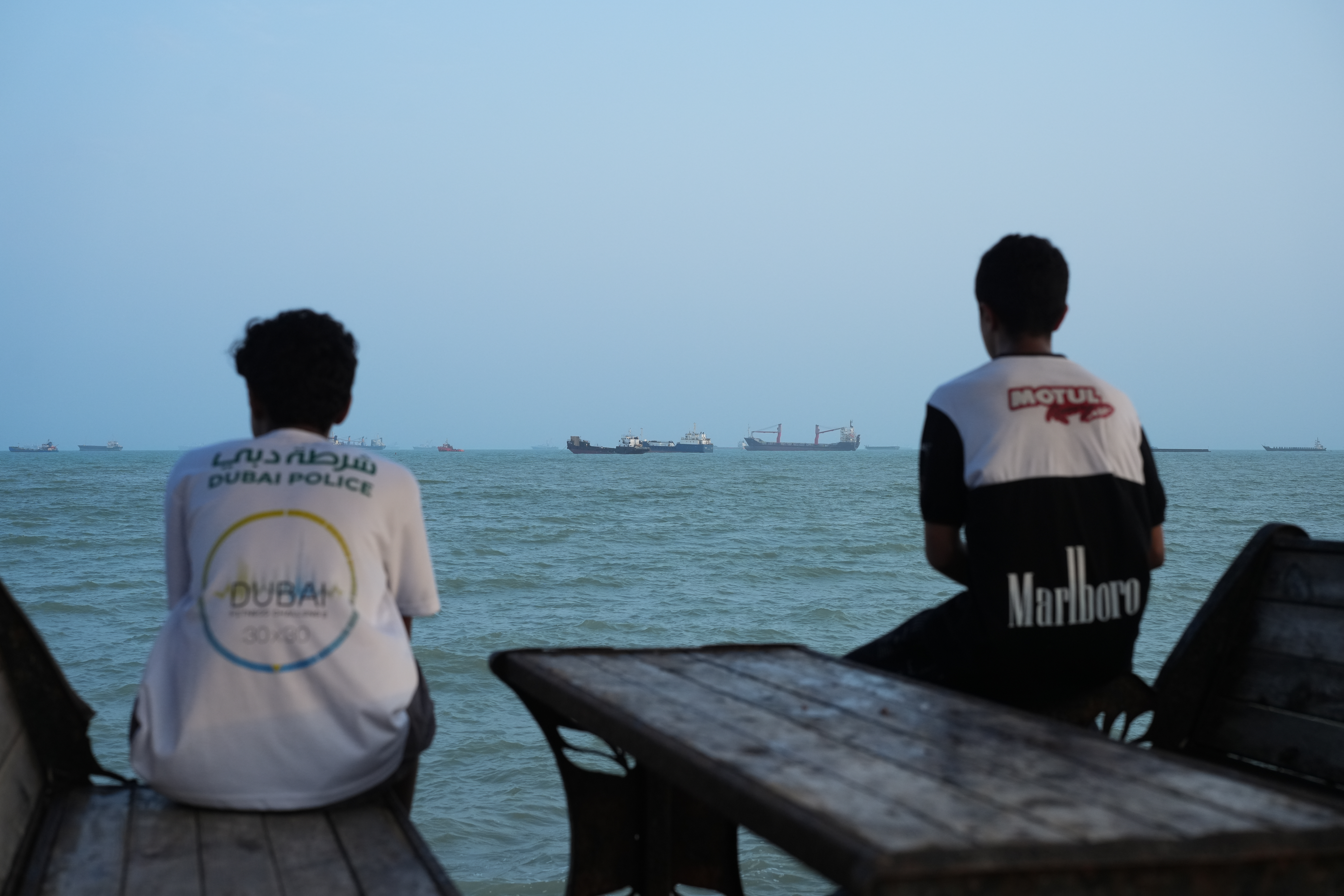 Two youth sit on the shoreline as ships are seen anchored at sea.