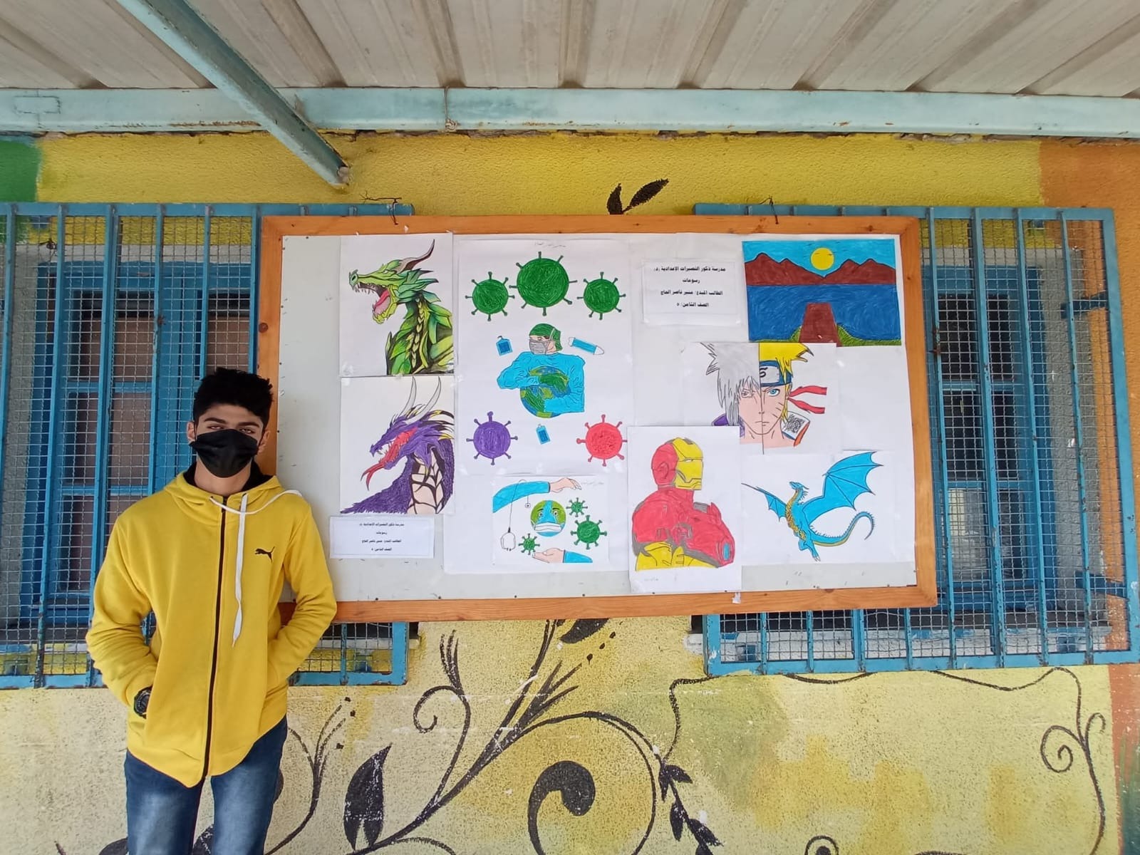 Muneer al-Haj standing next to an exhibition board featuring his artwork at his school before the war