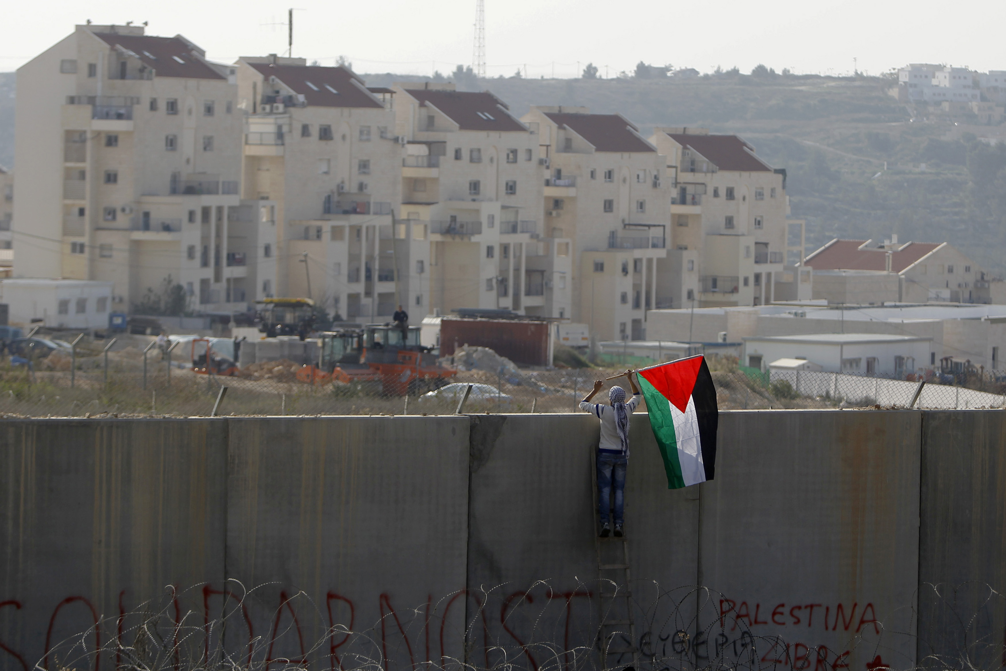 A man places the Palestinian flag on the controversial Israeli separation wall, with Israeli settlements showing in the background. Source: [REUTERS/Ammar Awad]