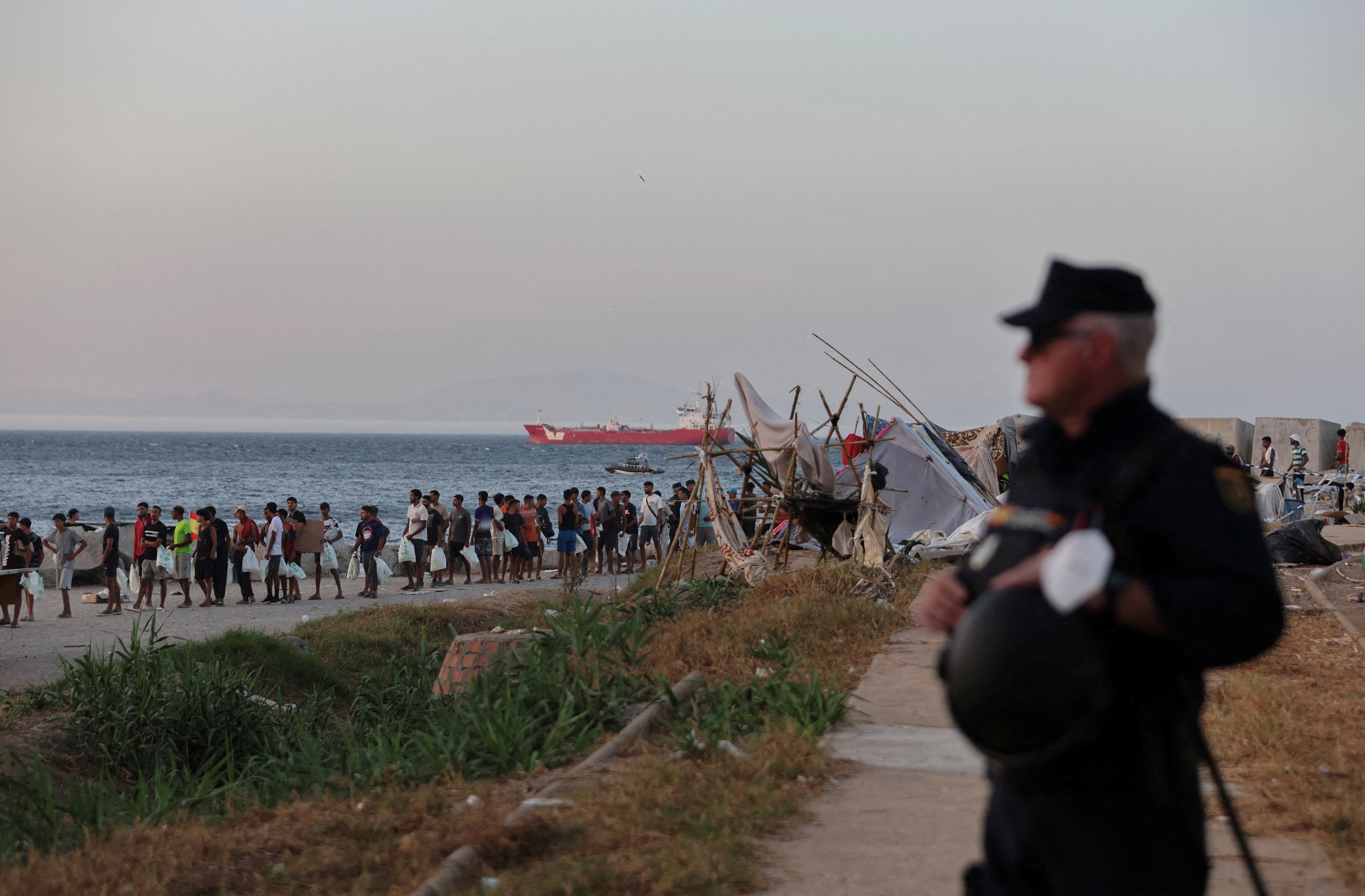 Migrants queue for food as a uniformed man looks on.