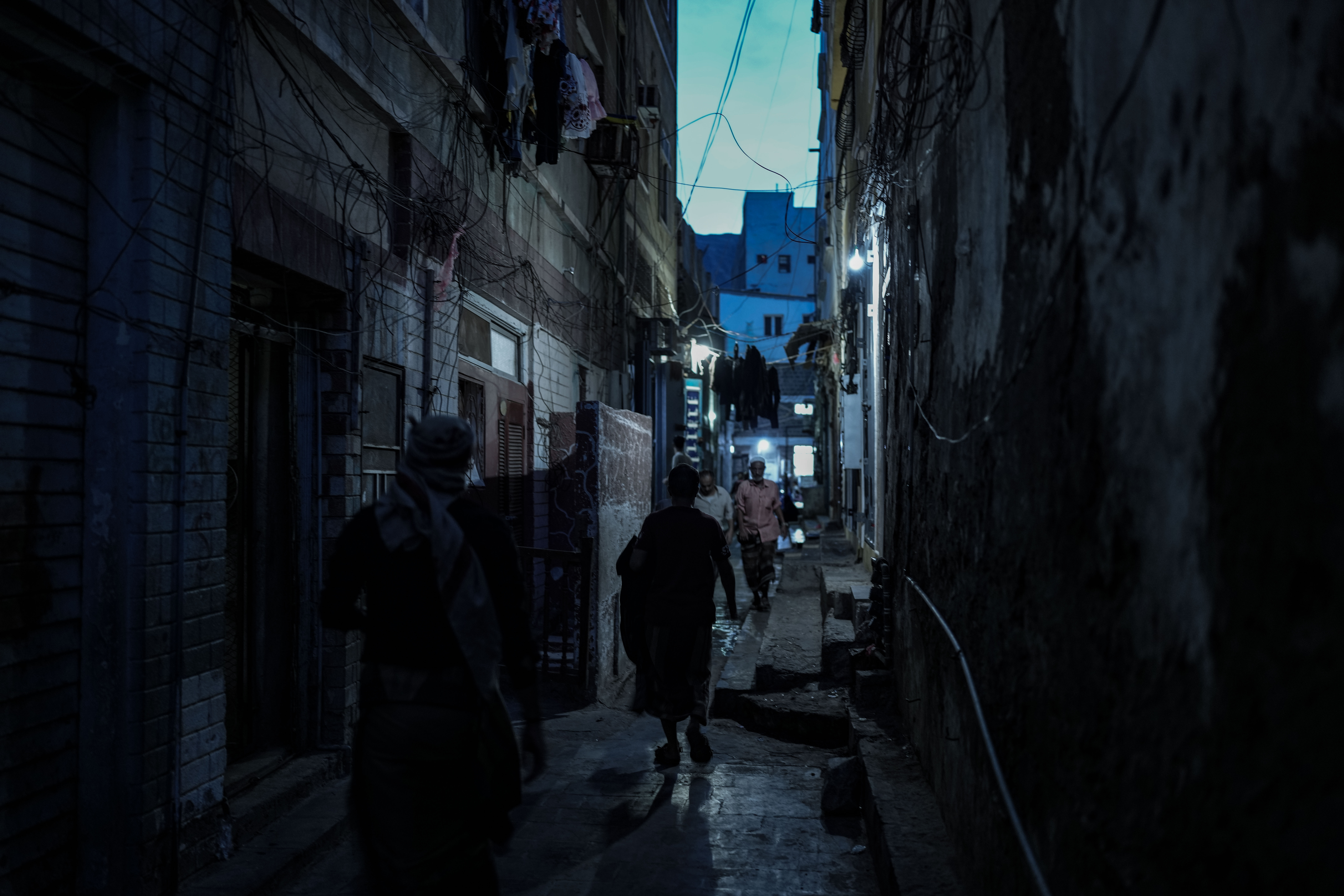 A resident walks through a dimly lit alley in Aden at night. During prolonged outages, light from shops, batteries and small alternative power sources becomes one of the few sources of illumination in some neighborhoods.